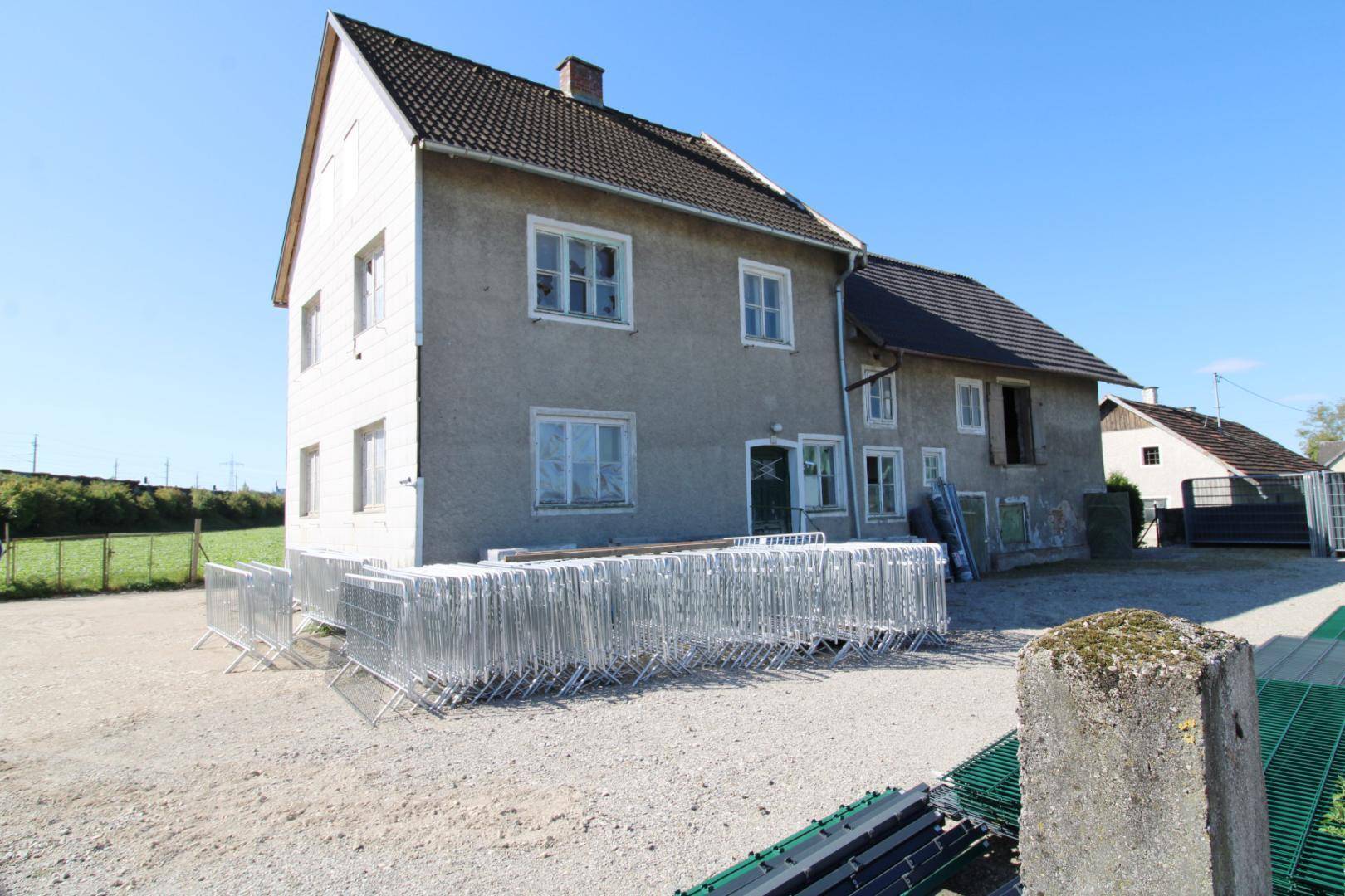 Traditionelles Wohnhaus mit grauer Fassade und Satteldach, umgeben von einem Schotterplatz und Metallzäunen.