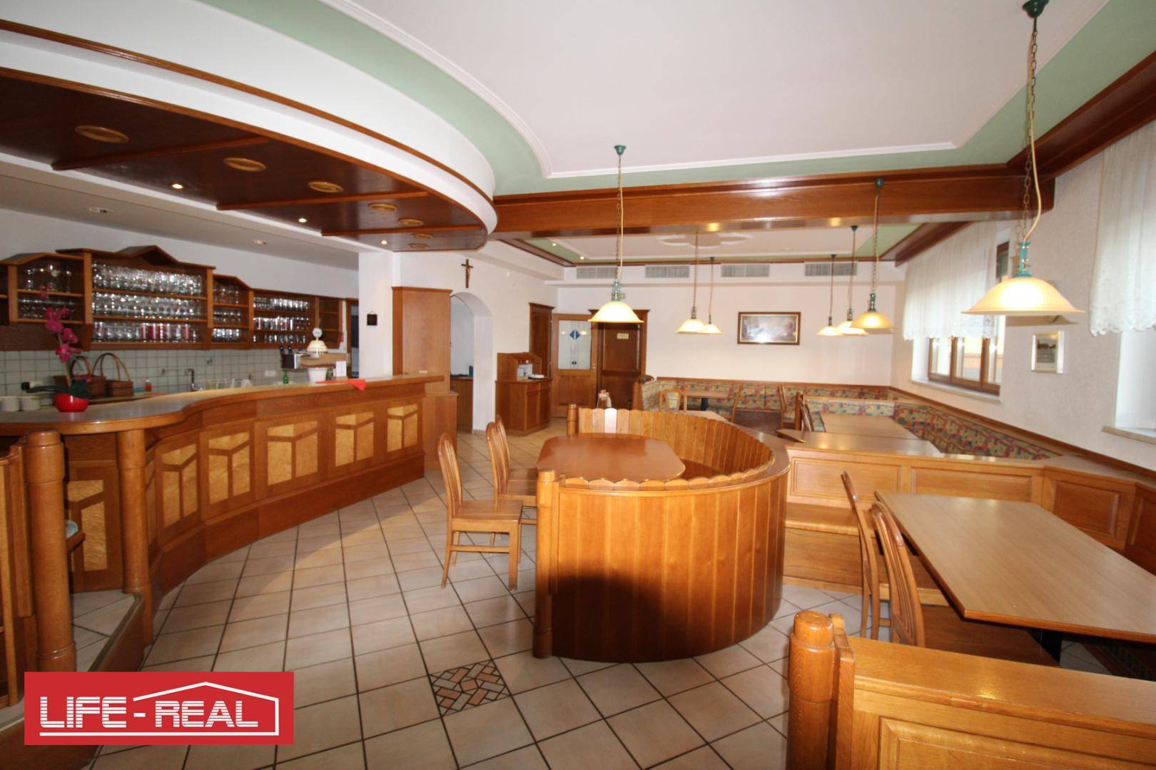 Weiterer Blick auf den Restaurantbereich mit geschwungener Holztheke, Sitzgelegenheiten und dekorativer Deckenbeleuchtung.