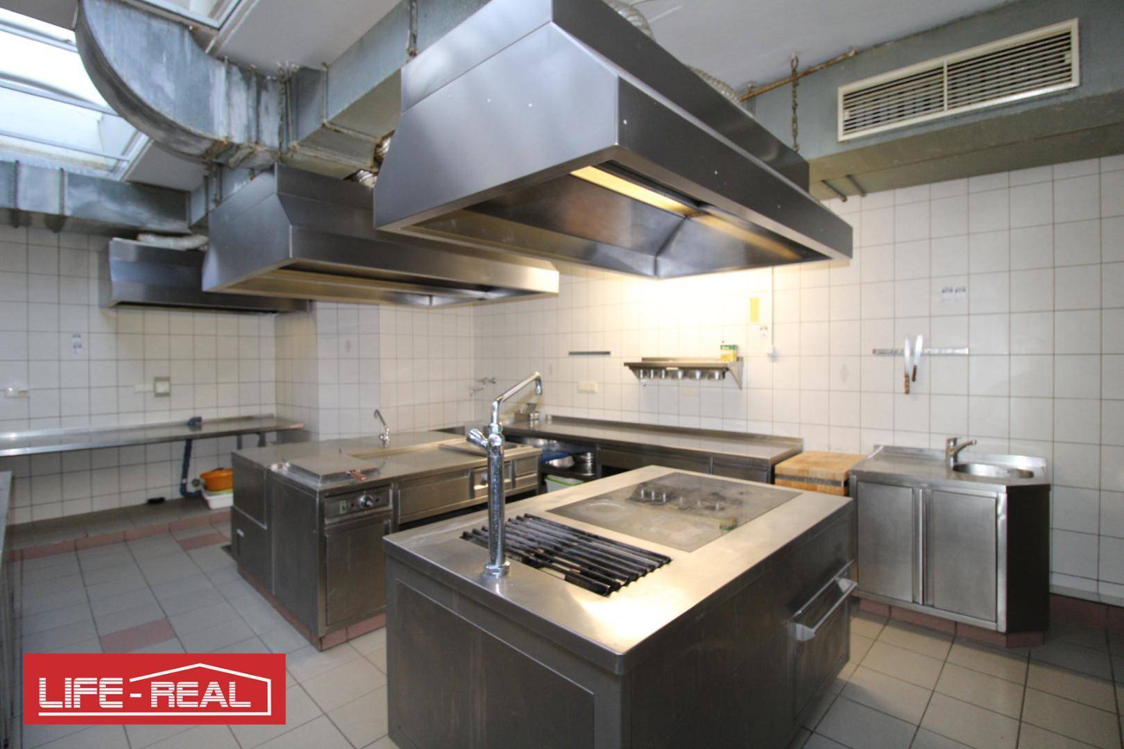 Professionelle Großküche mit Edelstahlgeräten, großen Dunstabzugshauben und gefliesten Wänden für den Gastronomiebetrieb.