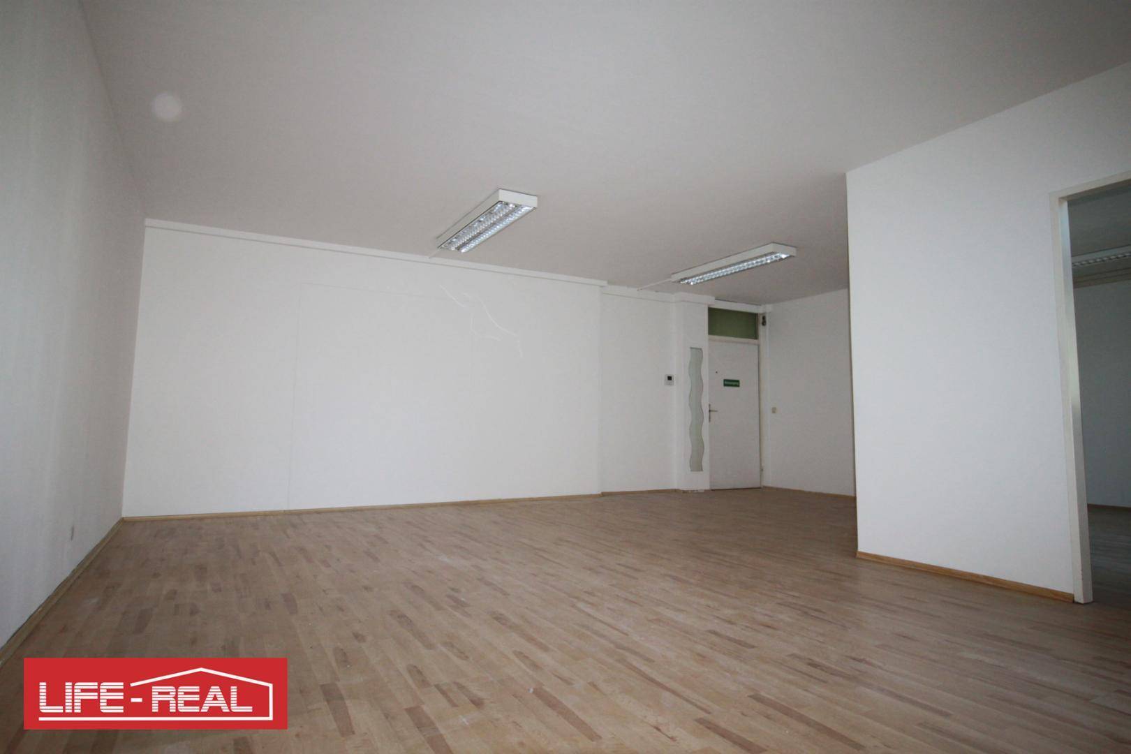 Geräumiger, leerer Büroraum mit hellem Laminatboden und einer Eingangstür.
