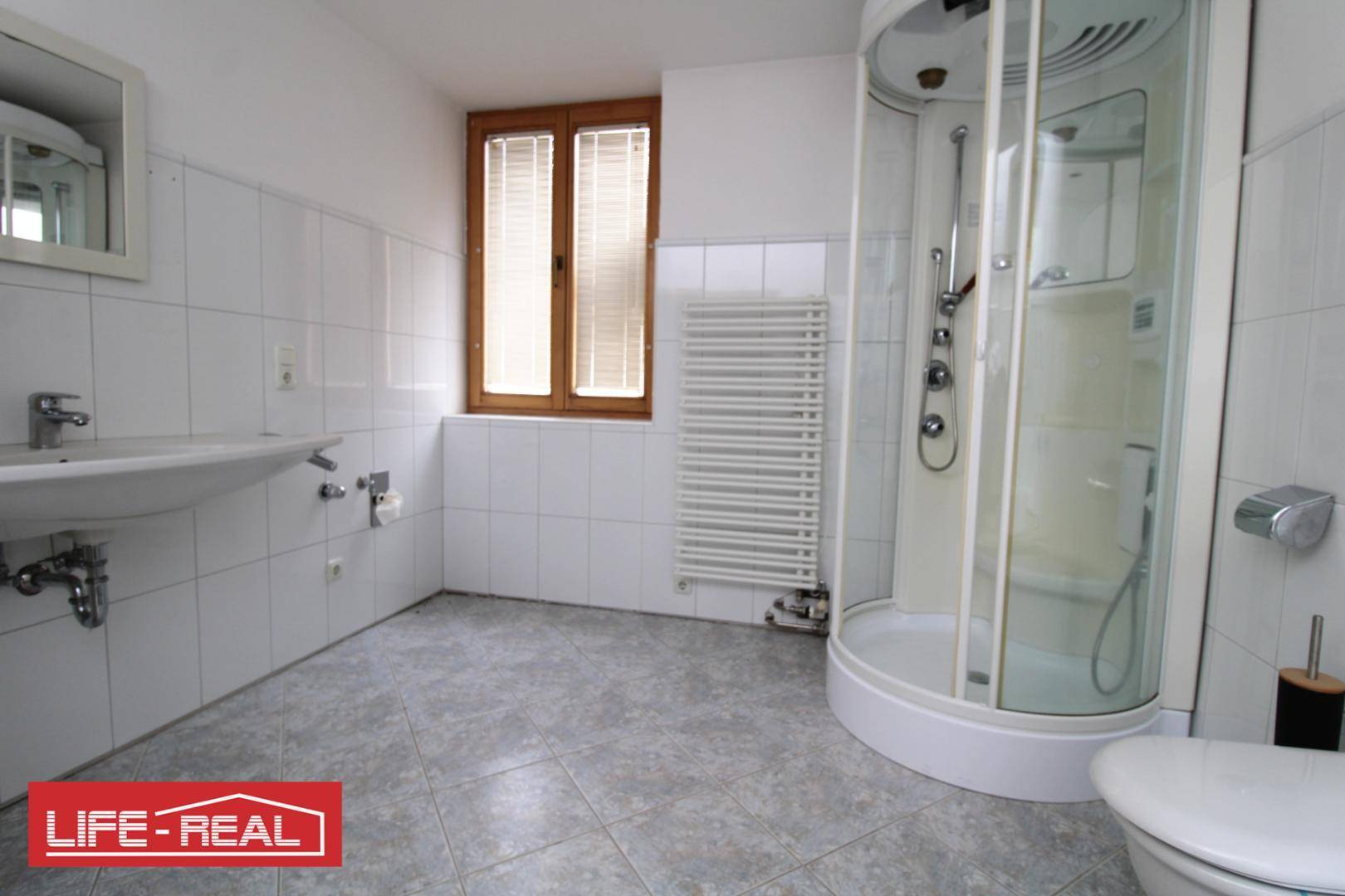 Helles Badezimmer mit weißen Fliesen, Duschkabine, Waschbecken und Fenster für Tageslicht.