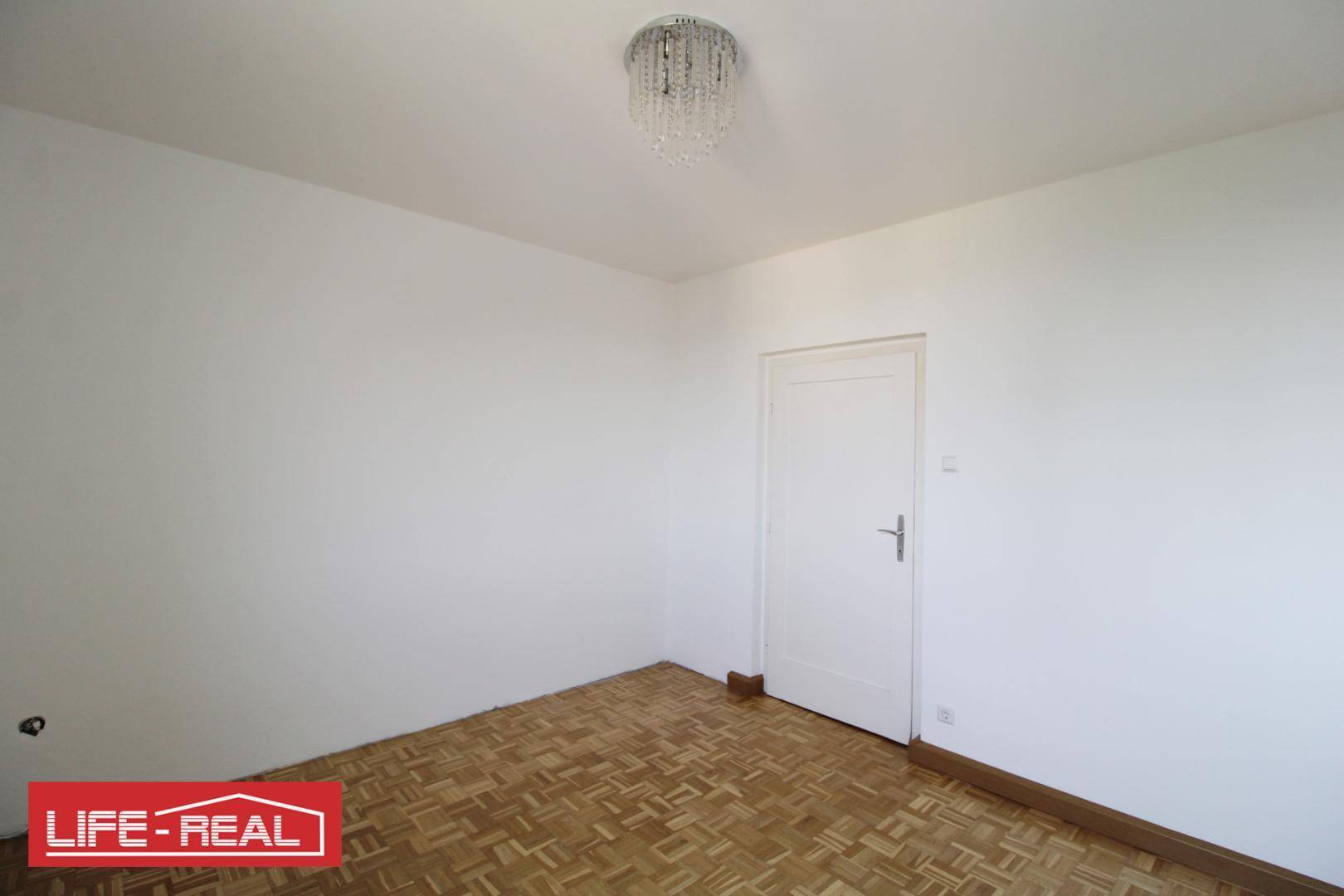 Leeres Zimmer mit Parkettboden, weißen Wänden und einem eleganten Kronleuchter.