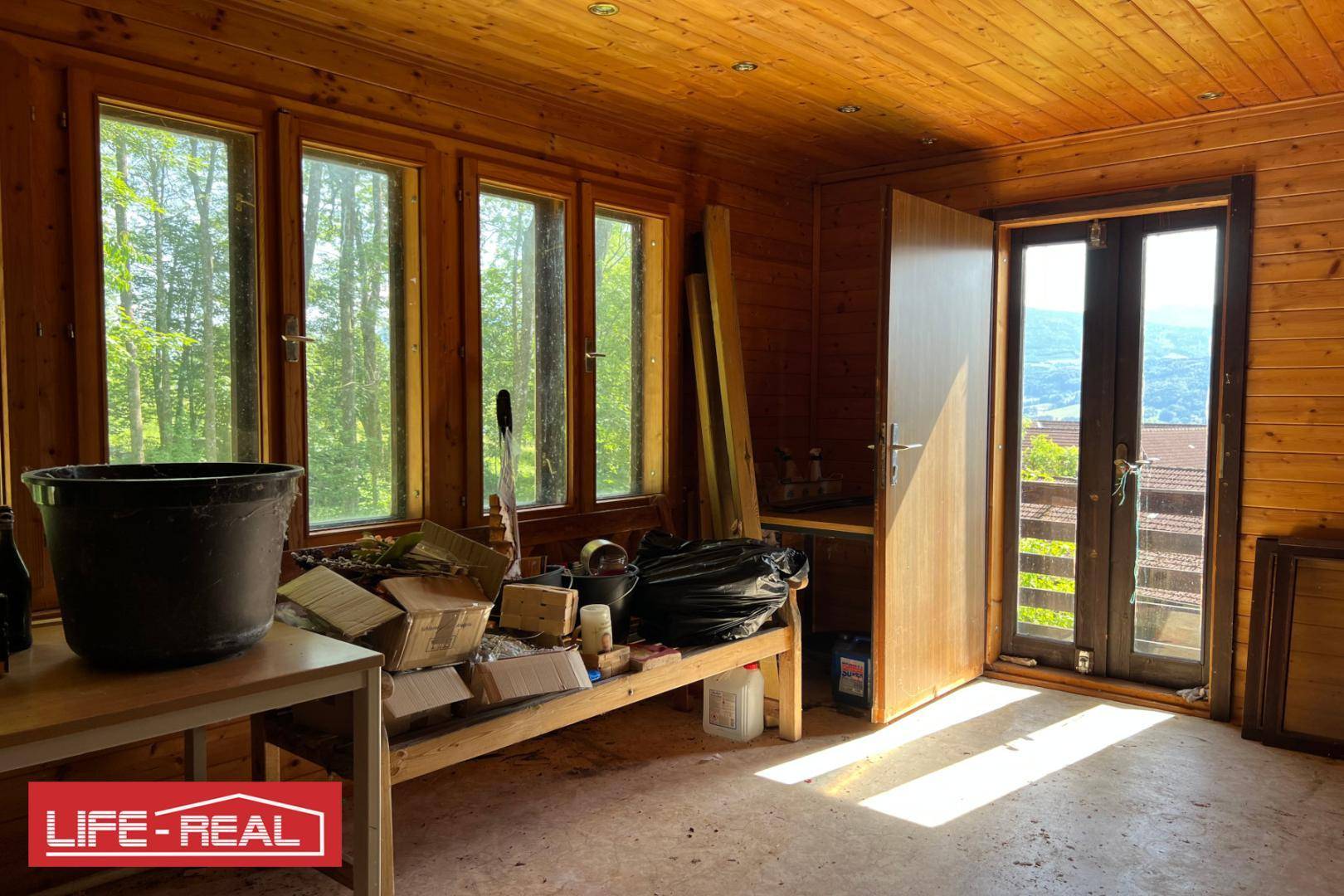Heller Raum mit Holzverkleidung an Wänden und Decke, großen Fenstern und Blick ins Grüne.