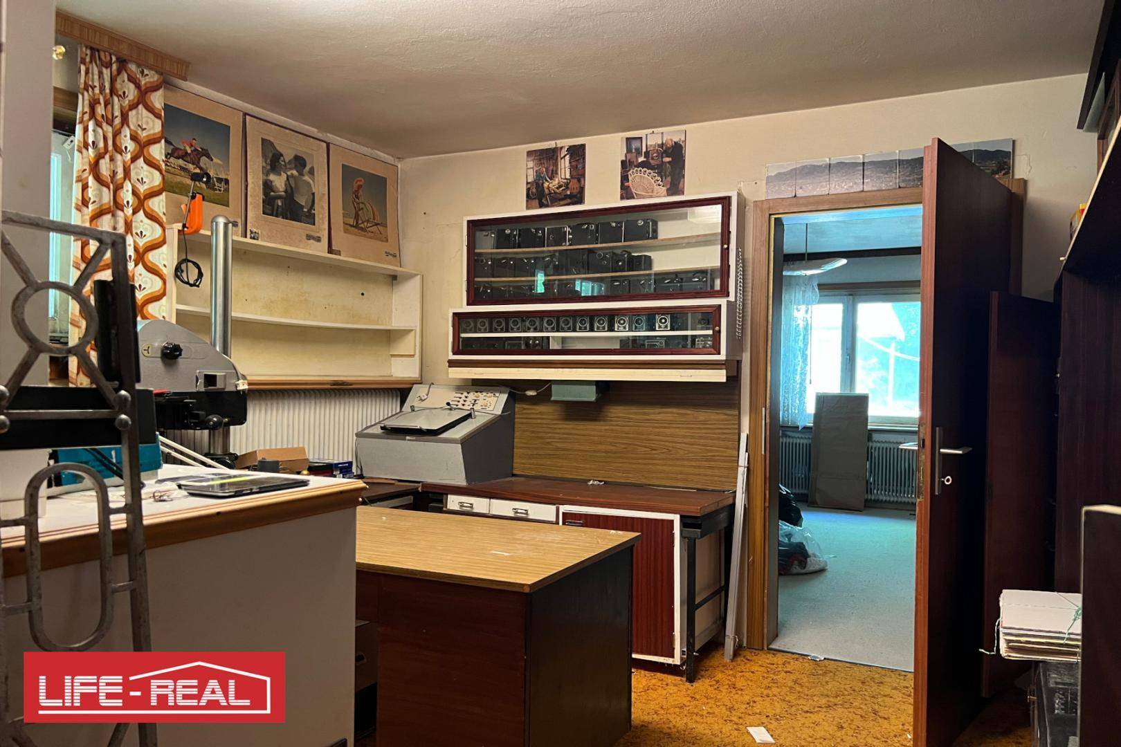 Abstellraum oder ehemaliges Büro mit verschiedenen Einrichtungsgegenständen und Durchgang zu weiterem Raum.