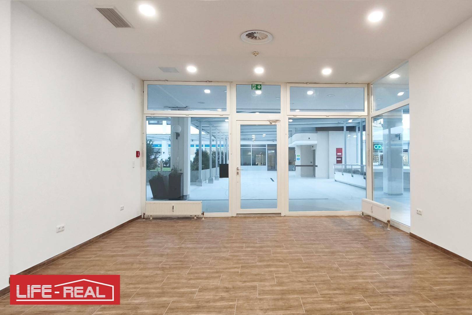 Heller, leerer Geschäftsraum mit großen Glasfronten und Heizkörpern unter den Fenstern.