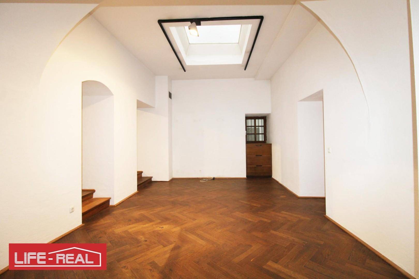Heller Raum mit Fischgrätparkett, einem großen Oberlicht und einer kleinen Nische mit Fenster, die zum Treppenaufgang führt.