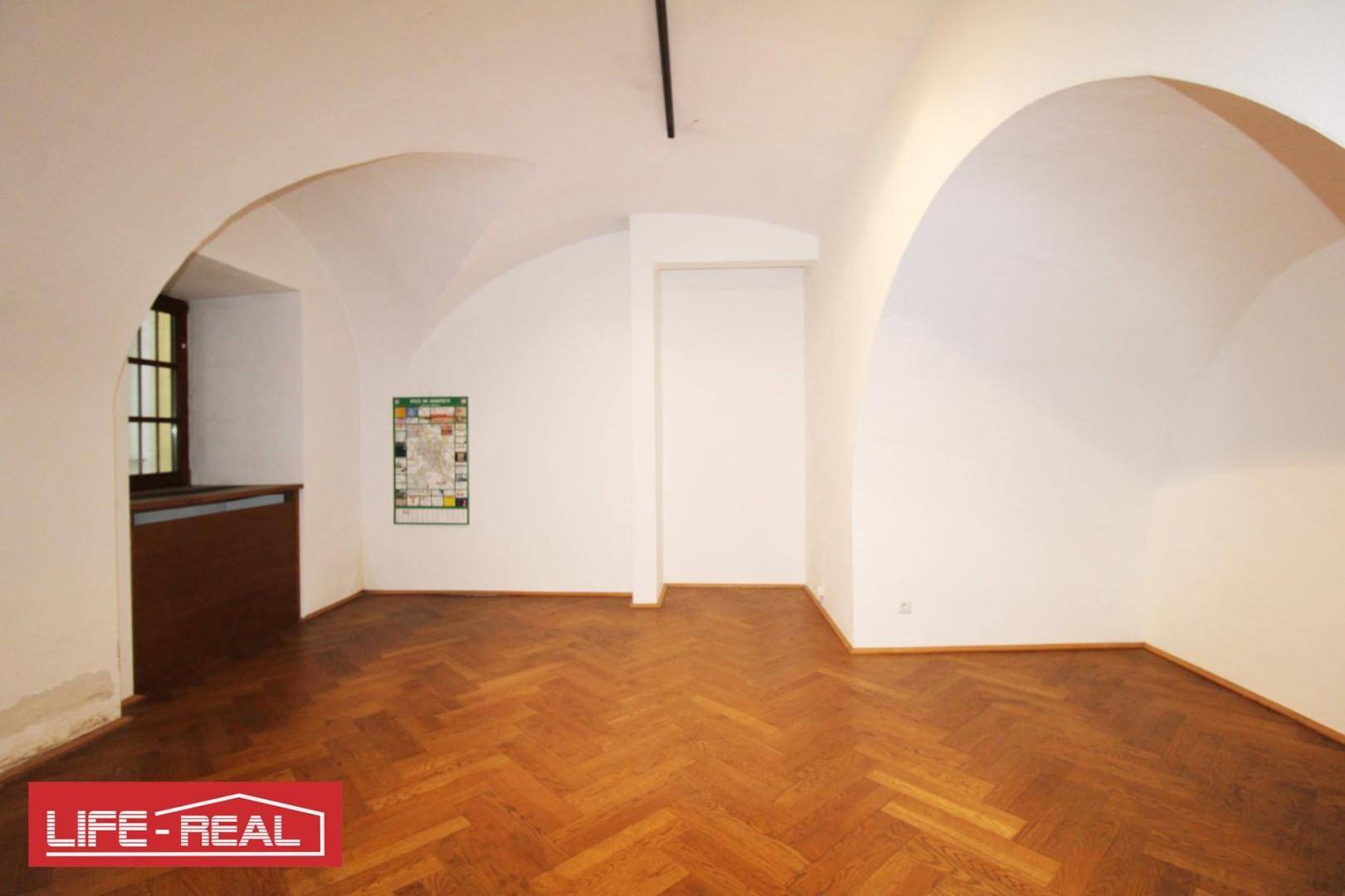 Geräumiger Innenraum mit Gewölbedecke und Fischgrätparkett, ergänzt durch ein kleines Fenster und eine schlichte Wandgestaltung.