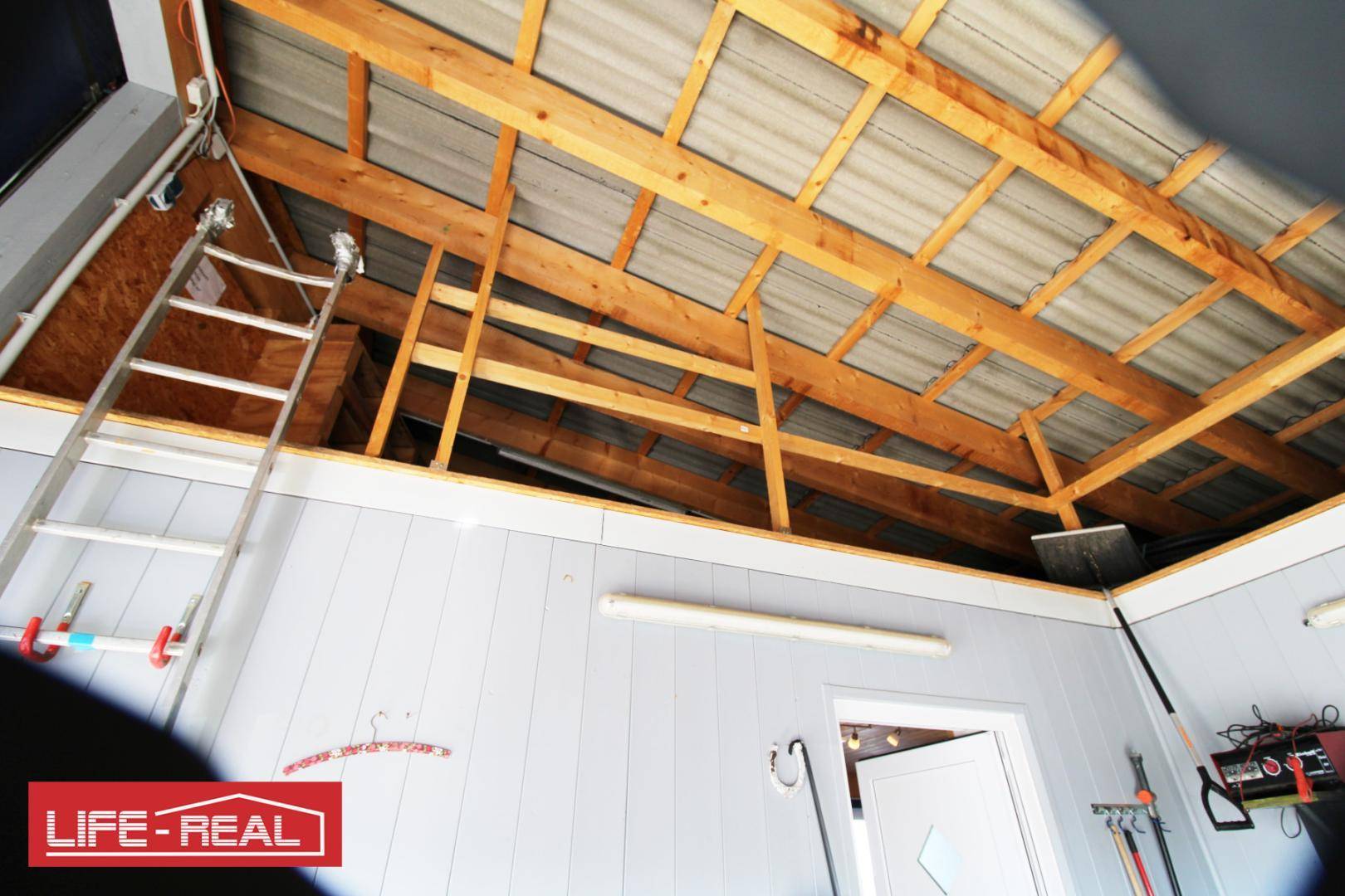 Werkstatt oder Lagerraum mit einer Leiter, Werkzeugen und einer sichtbaren Dachkonstruktion aus Holz und Wellblech.