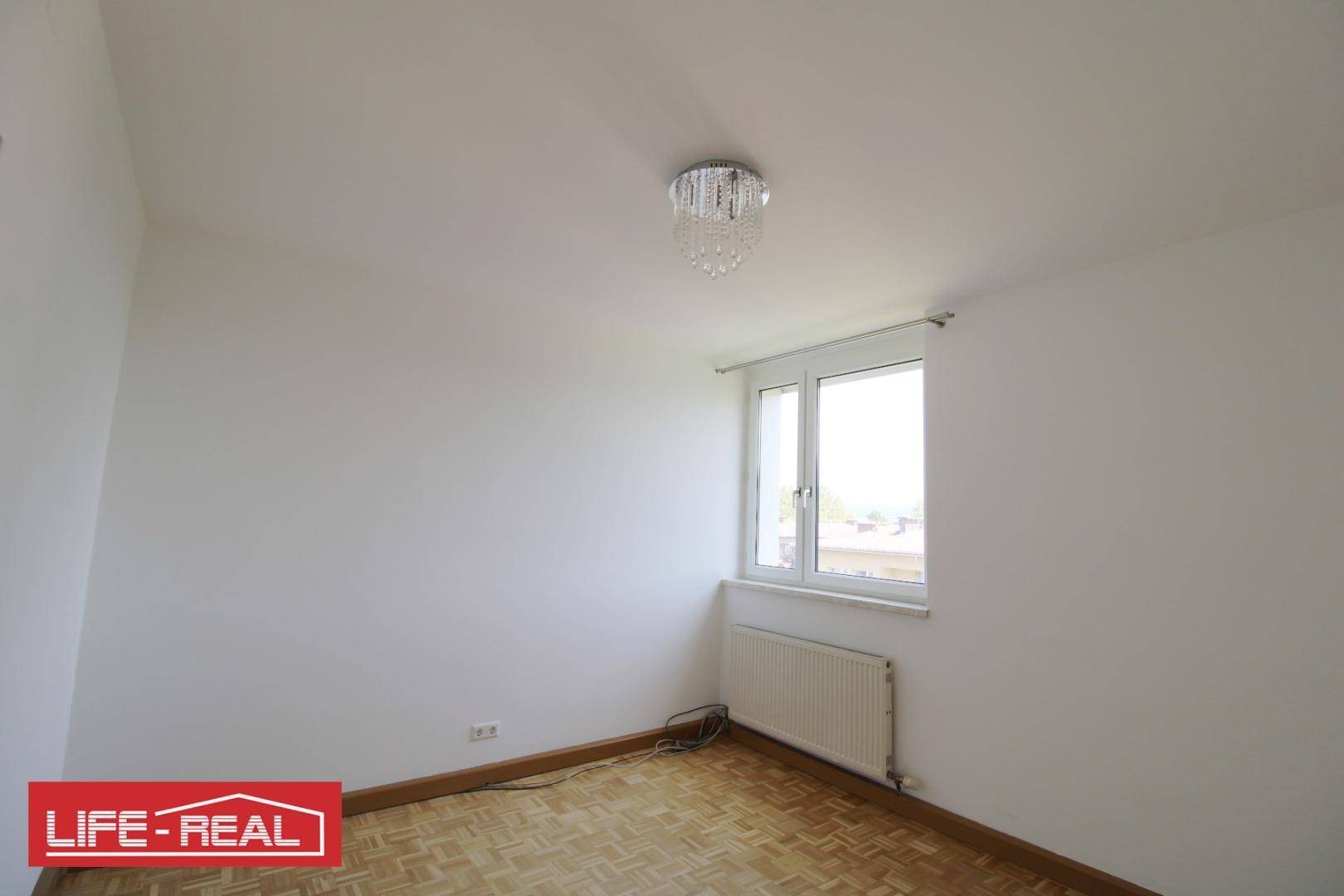 Helles Zimmer mit Parkettboden, Fenster und einem dekorativen Kronleuchter.