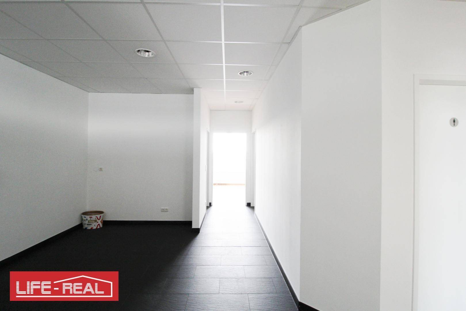 Langer, heller Flur in einem Bürogebäude mit dunklem Boden und weißen Wänden, führt zu einem weiteren Raum.