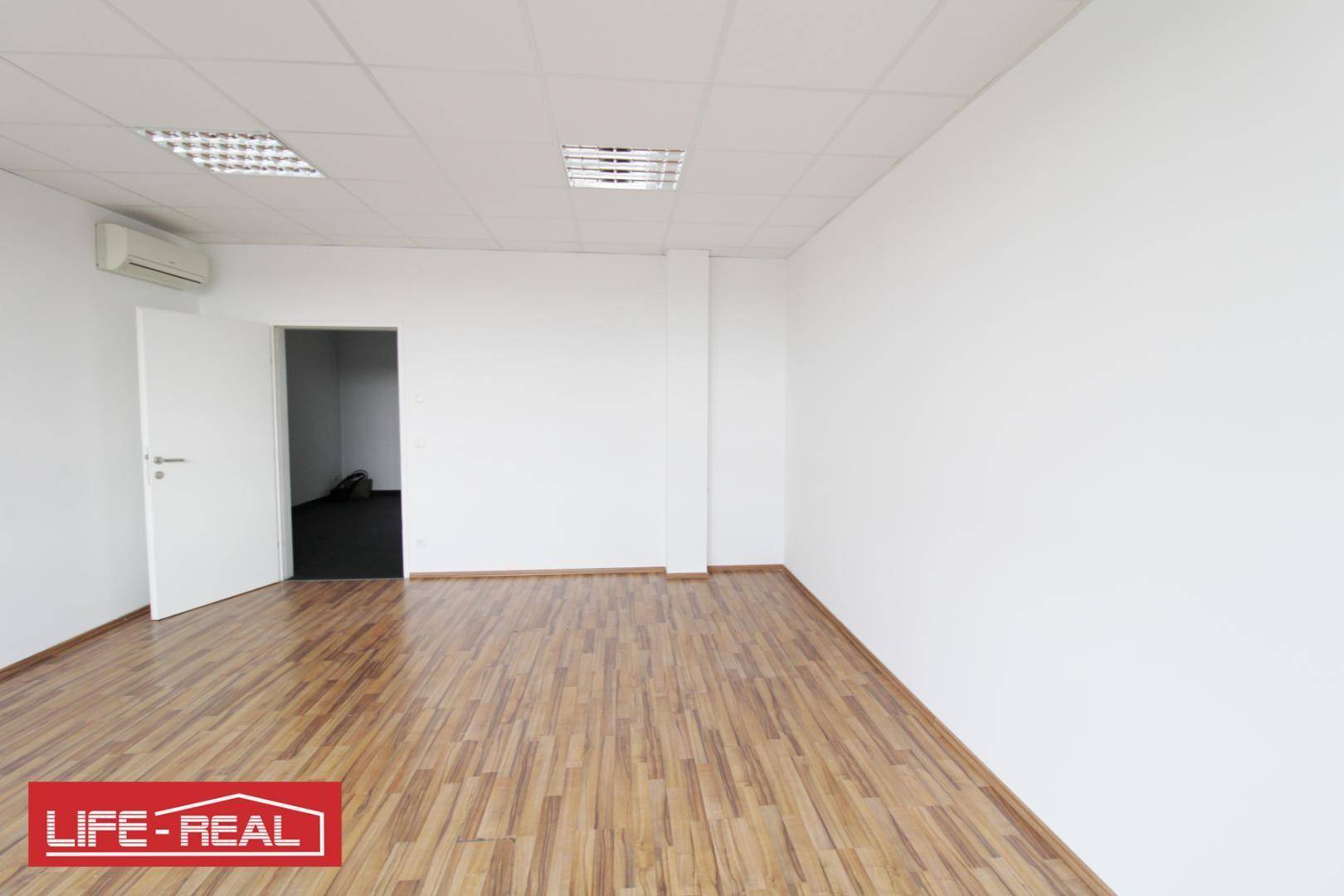 Leerer Büroraum mit hellem Laminatboden und weißen Wänden, eine offene Tür führt zu einem angrenzenden Bereich.
