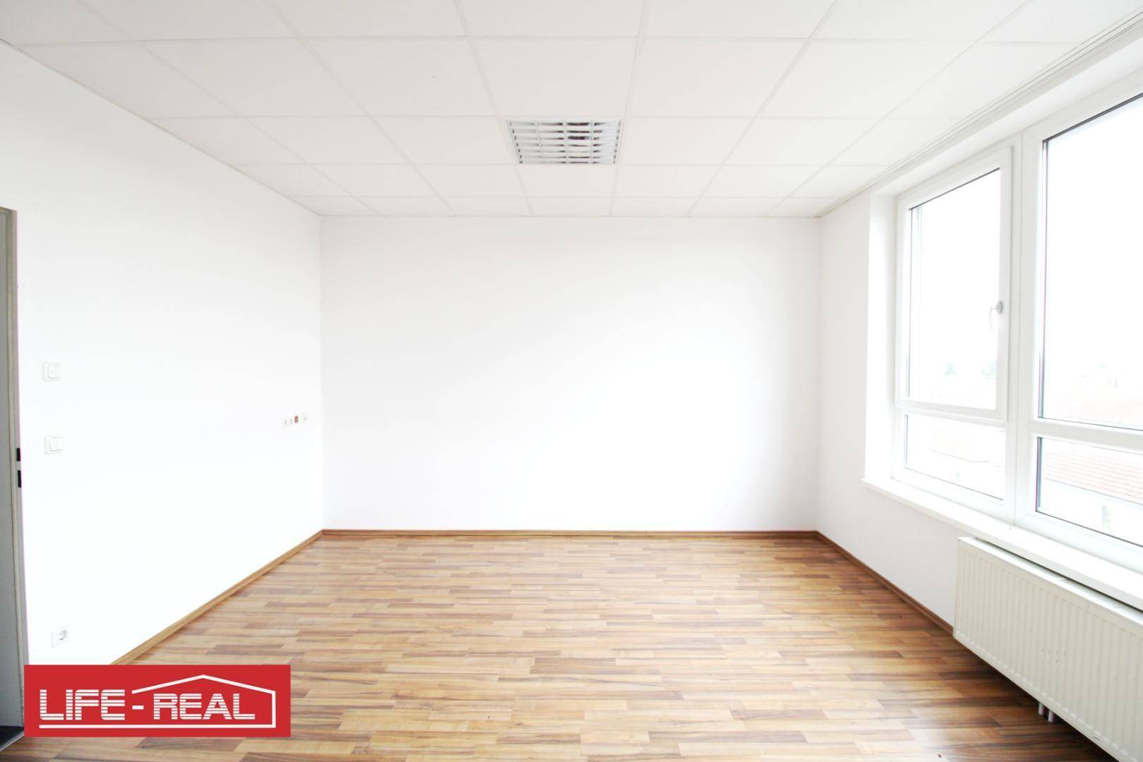 Leerer Büroraum mit hellem Laminatboden und weißen Wänden, ausgestattet mit einem großen Fenster und Heizkörper.