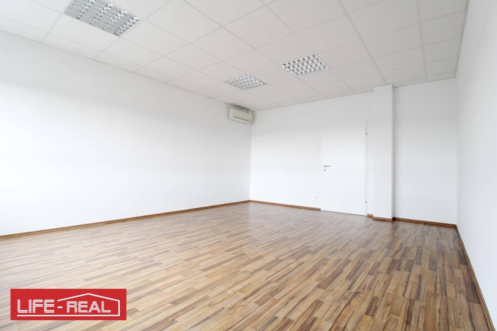 Geräumiger Büroraum mit hellem Laminatboden, weißen Wänden und einer Klimaanlage an der Wand.
