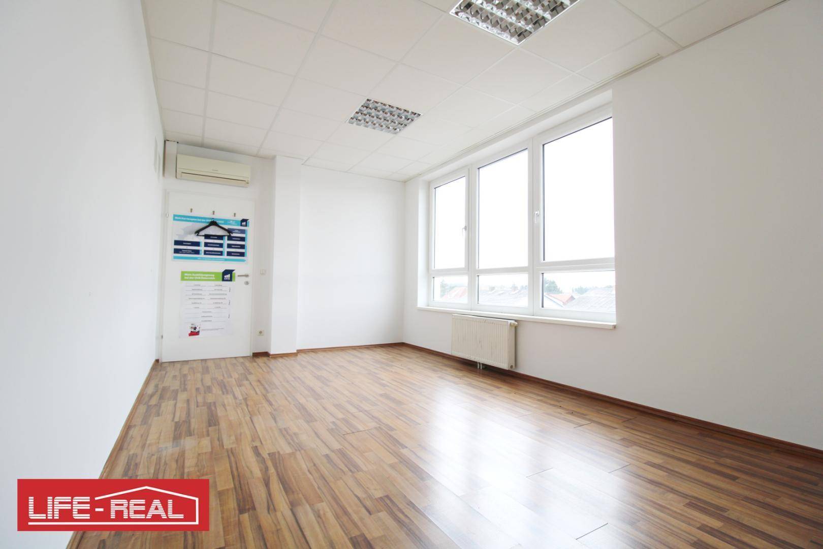 Helles Büro mit Laminatboden, weißen Wänden und einem großen Fenster, das viel Tageslicht hereinlässt.