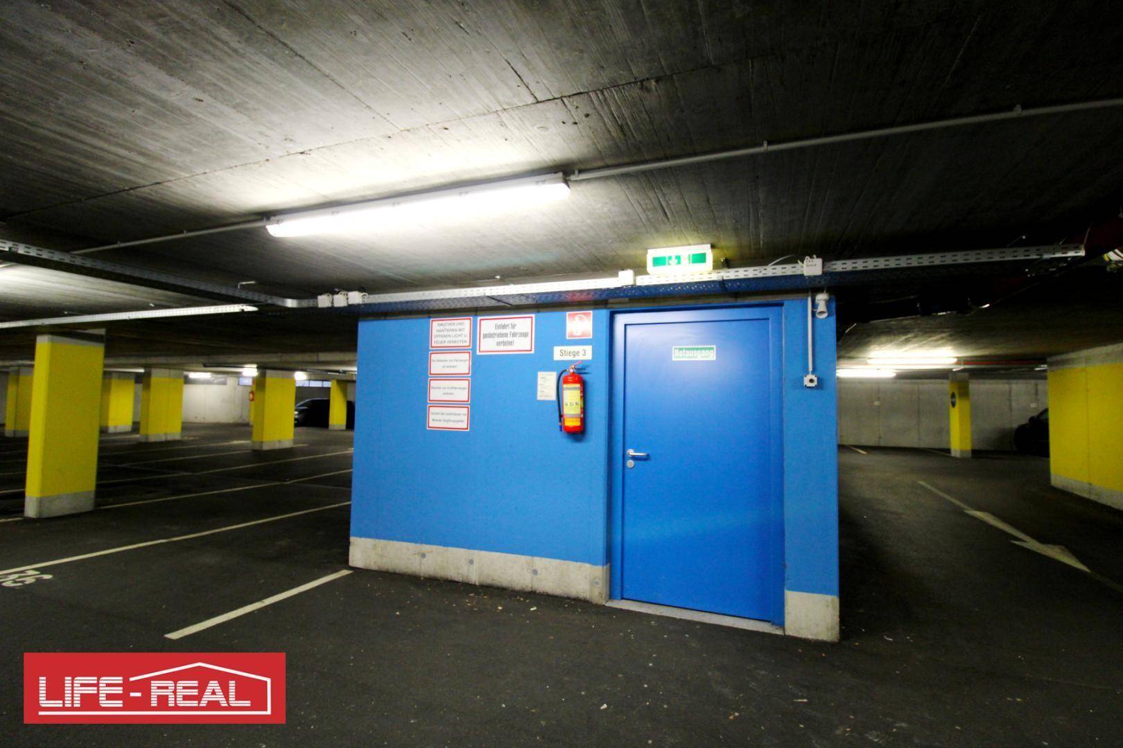 Tiefgarage mit Betonwänden und gelben Säulen, ein blauer Raum mit Tür und Feuerlöscher ist sichtbar.