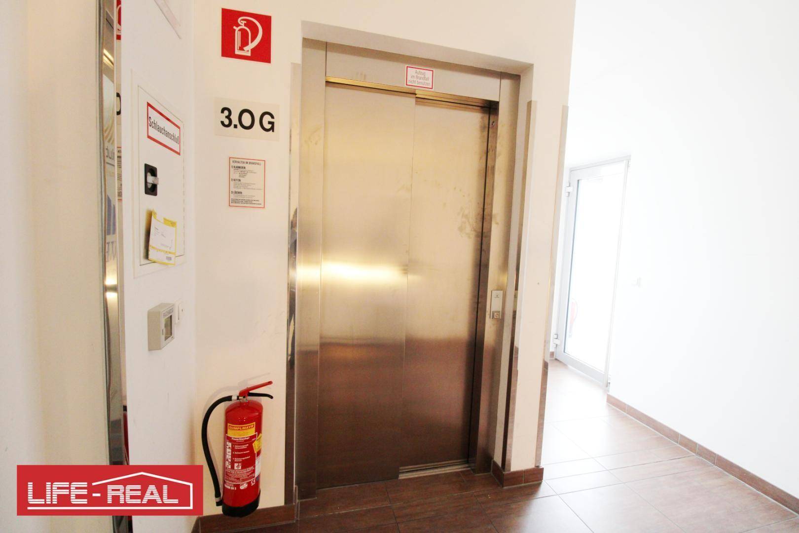 Moderner Aufzug mit Edelstahltüren in einem hellen Flur, daneben ein Feuerlöscher für die Sicherheit.