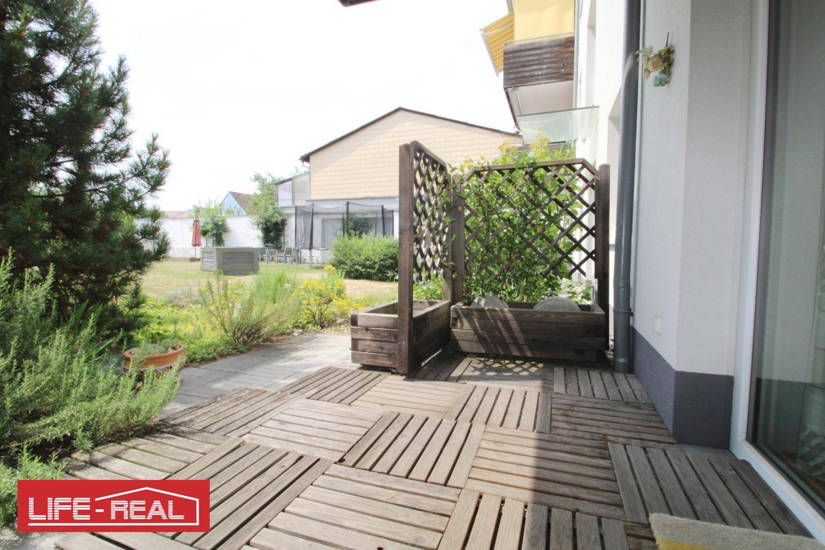 Geräumige Terrasse mit Holzfliesen und Zugang zum Garten, umgeben von Grünflächen.