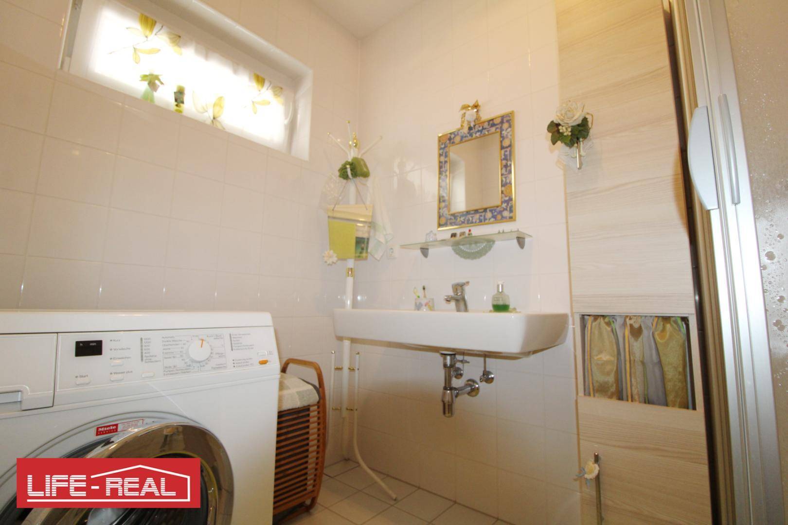 Helles Badezimmer mit Waschmaschine, Waschbecken und Duschkabine, gefliest in hellen Tönen.