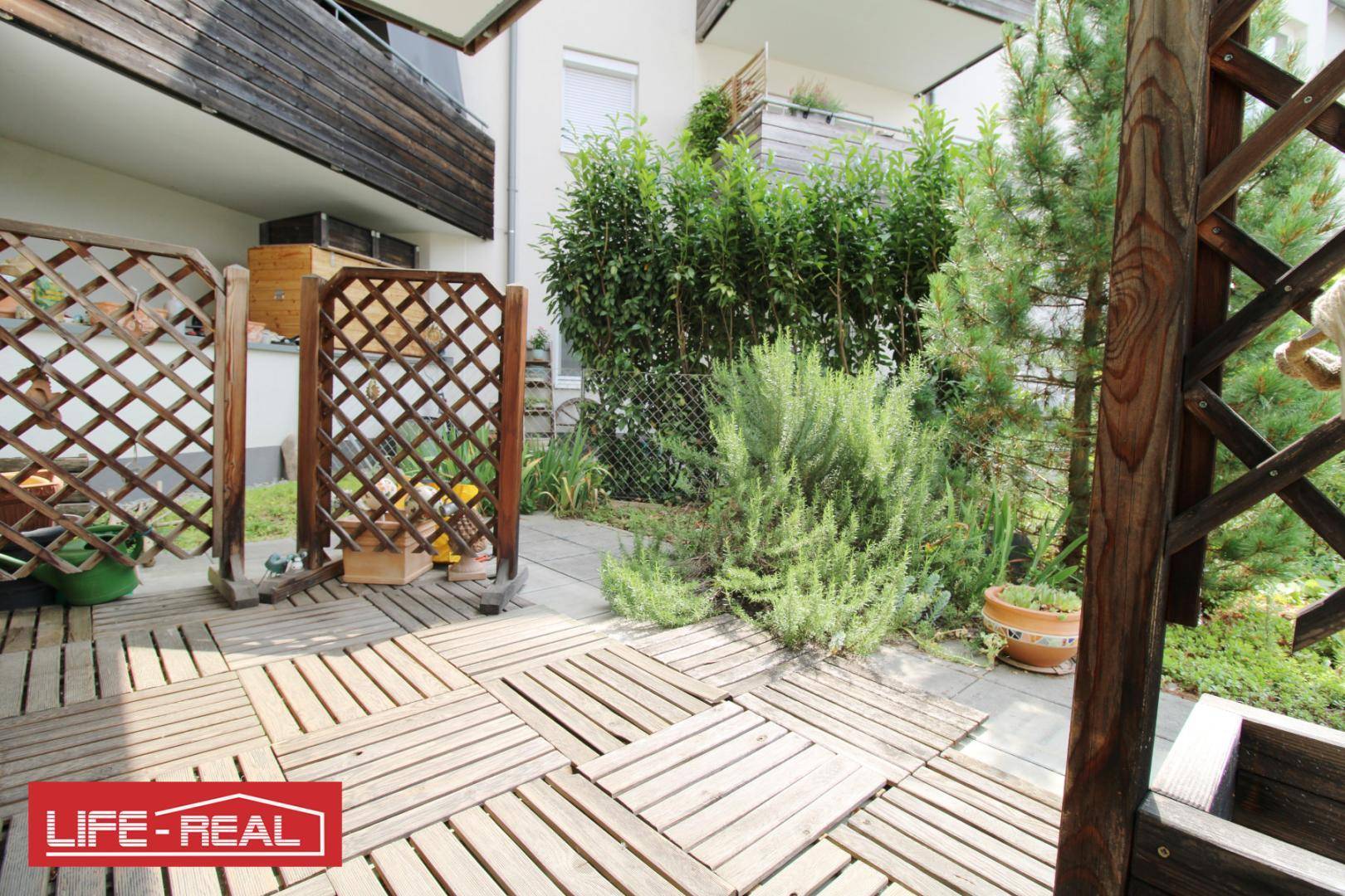 Sonniger Terrassenbereich mit Holzfliesen, umgeben von Pflanzen und Sichtschutz.