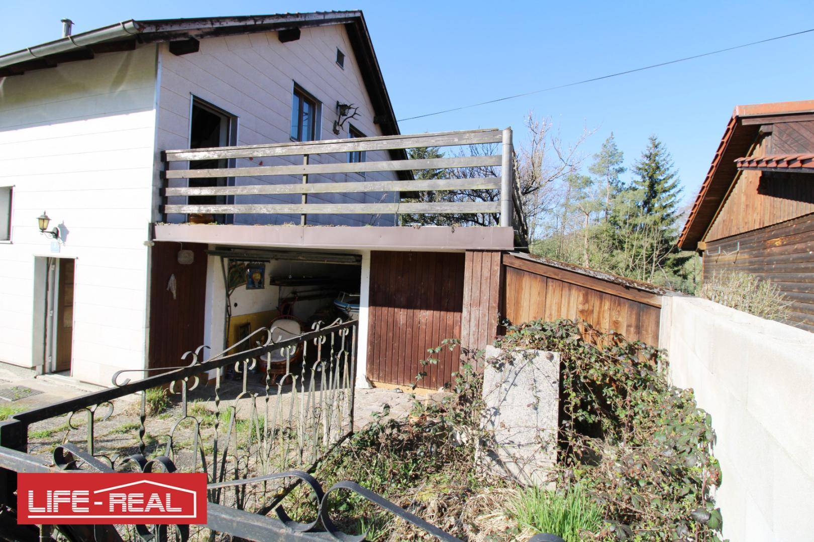 Hausfassade mit einem Holzbalkon über einer Garage und einem schmiedeeisernen Tor.