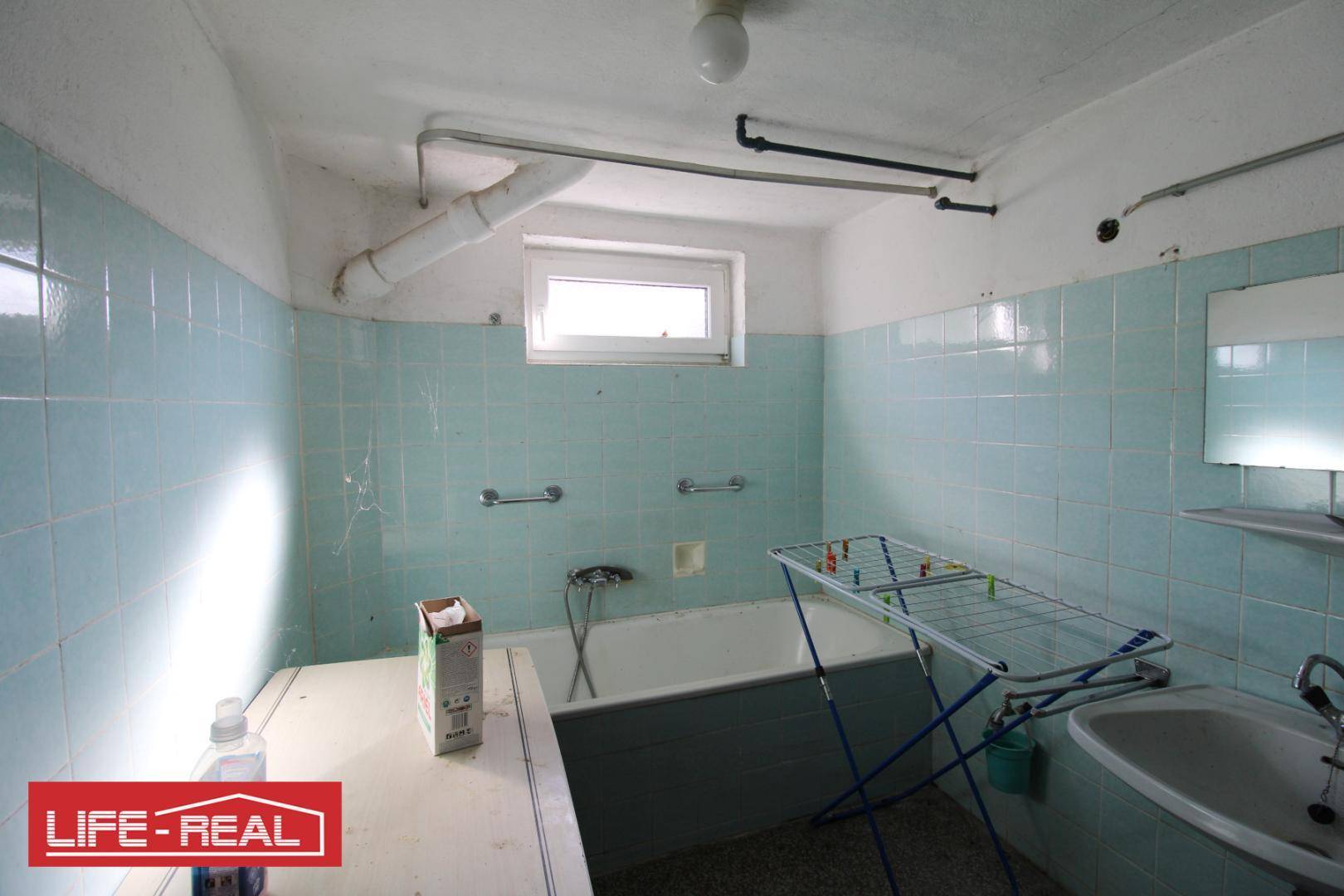 Badezimmer mit hellblauen Fliesen, Badewanne und Fenster, in einem älteren Zustand.