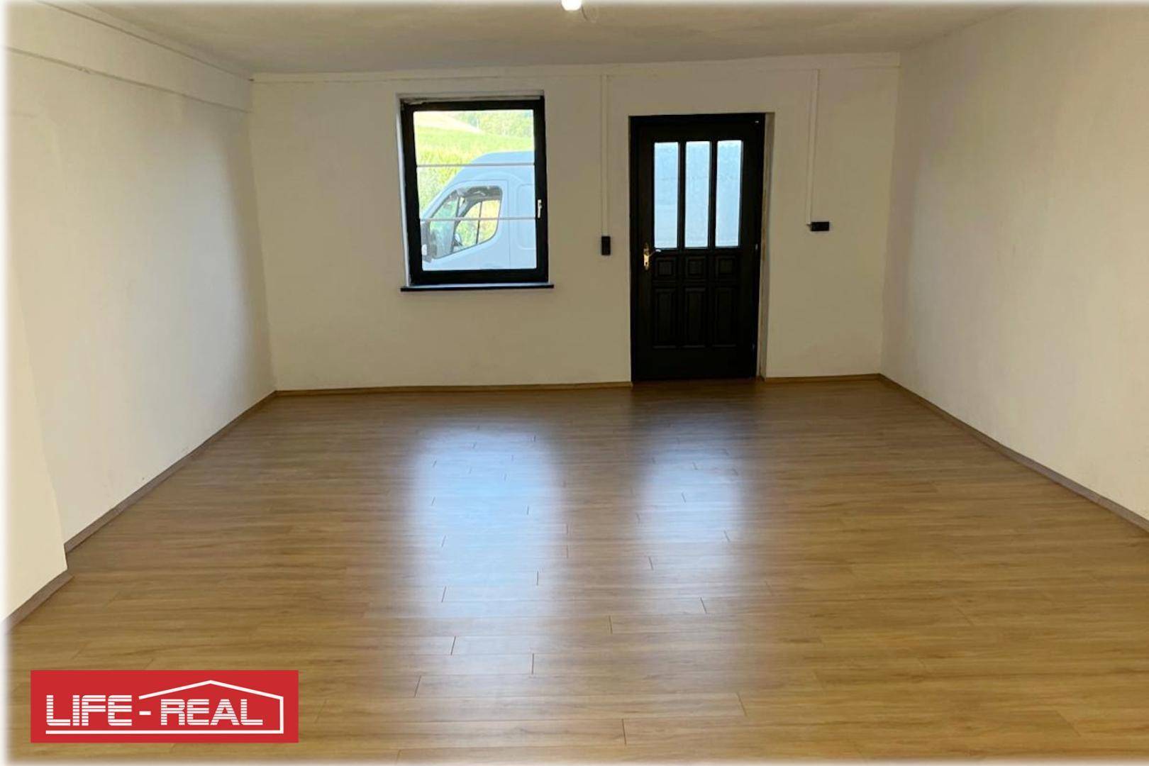 Heller, leerer Raum mit Laminatboden, weißen Wänden, einem Fenster und einer dunklen Tür.