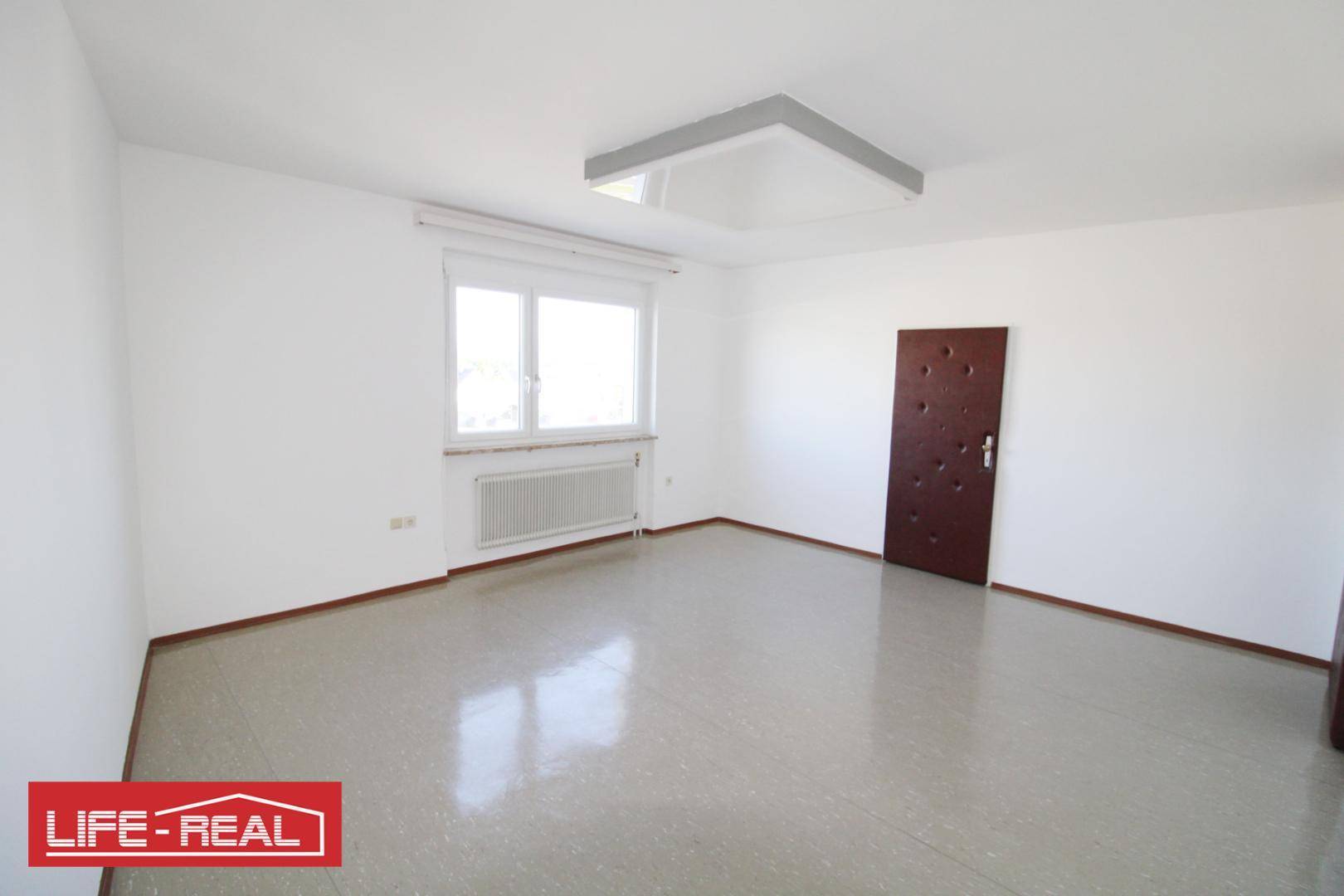 Heller, leerer Raum mit glänzendem Boden, einem Fenster, Deckenleuchte und einer dunklen Tür.
