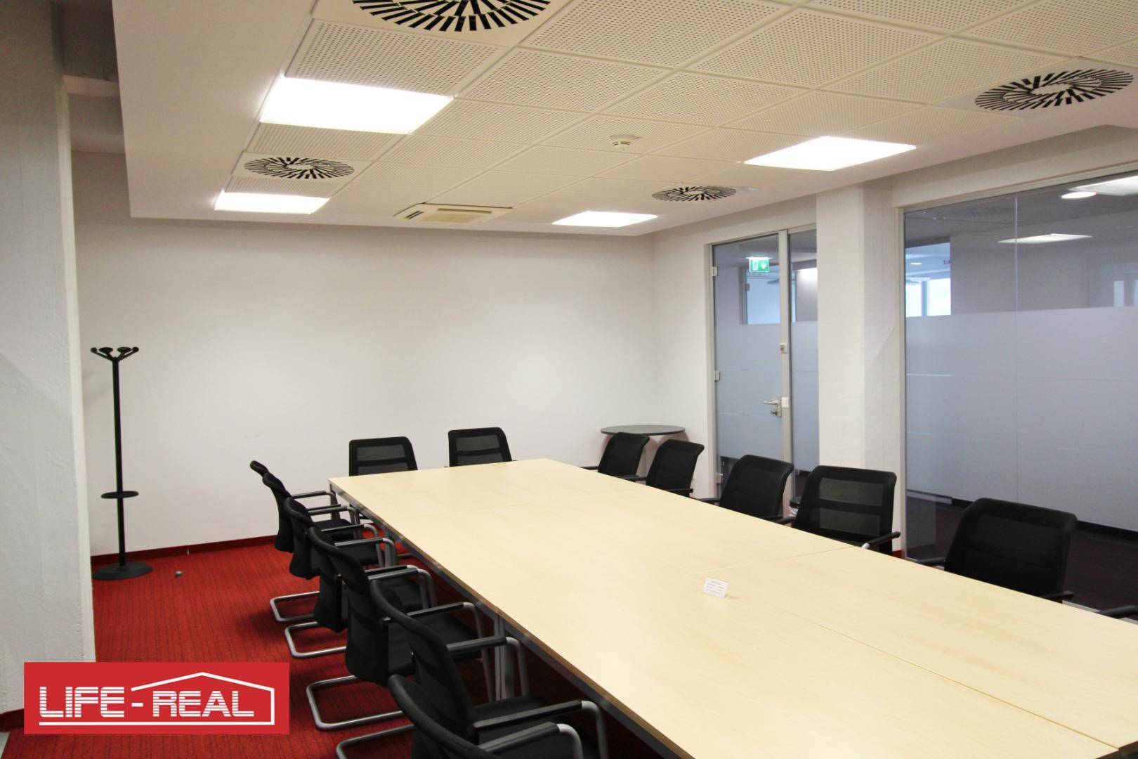 Gut ausgestatteter Konferenzraum mit hellem Holztisch, schwarzen Stühlen und rotem Teppich, ideal für Besprechungen.