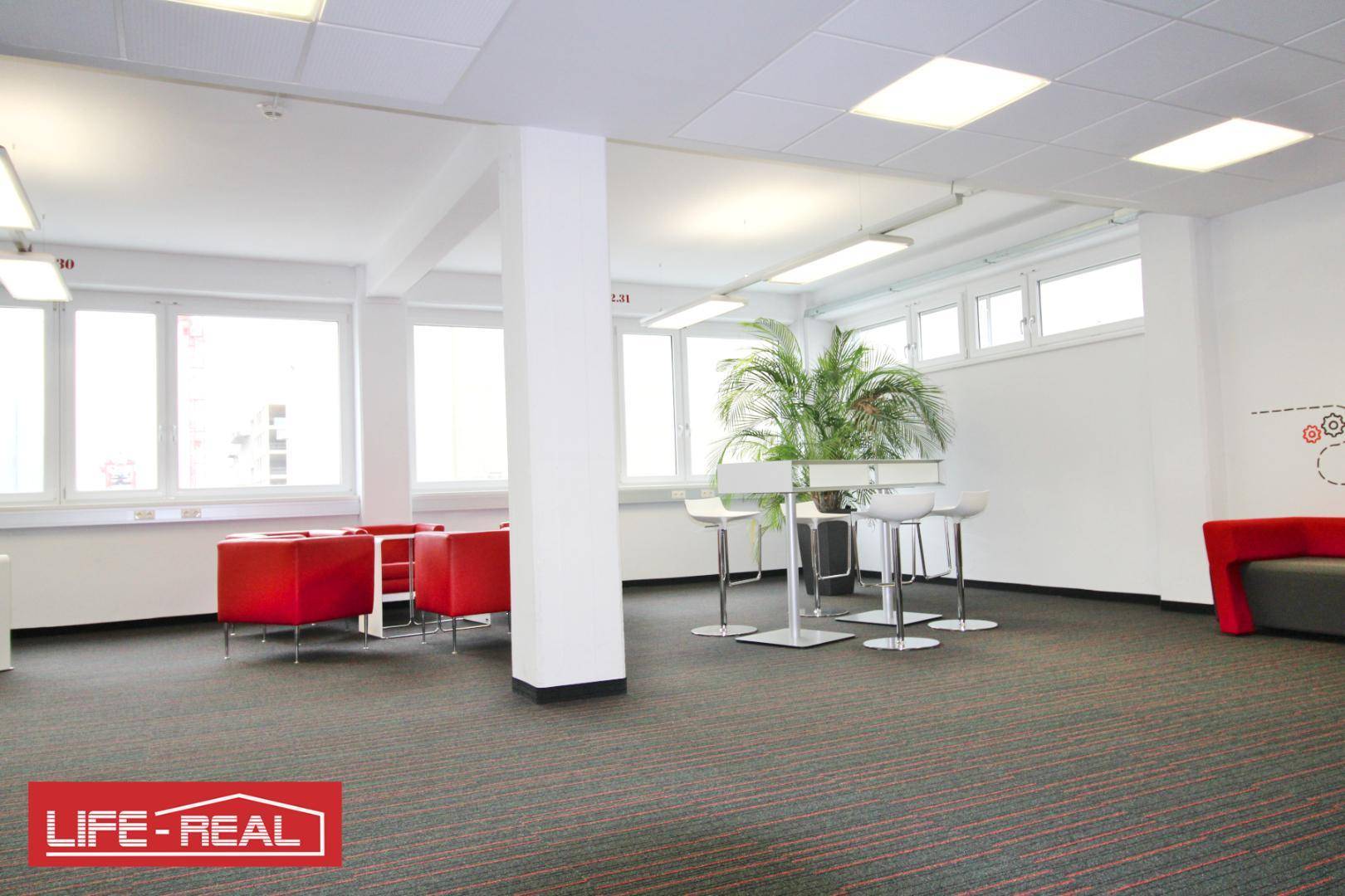 Gemütlicher Lounge-Bereich im Büro mit roten Sesseln, hohen Tischen und einer großen Pflanze vor den Fenstern.