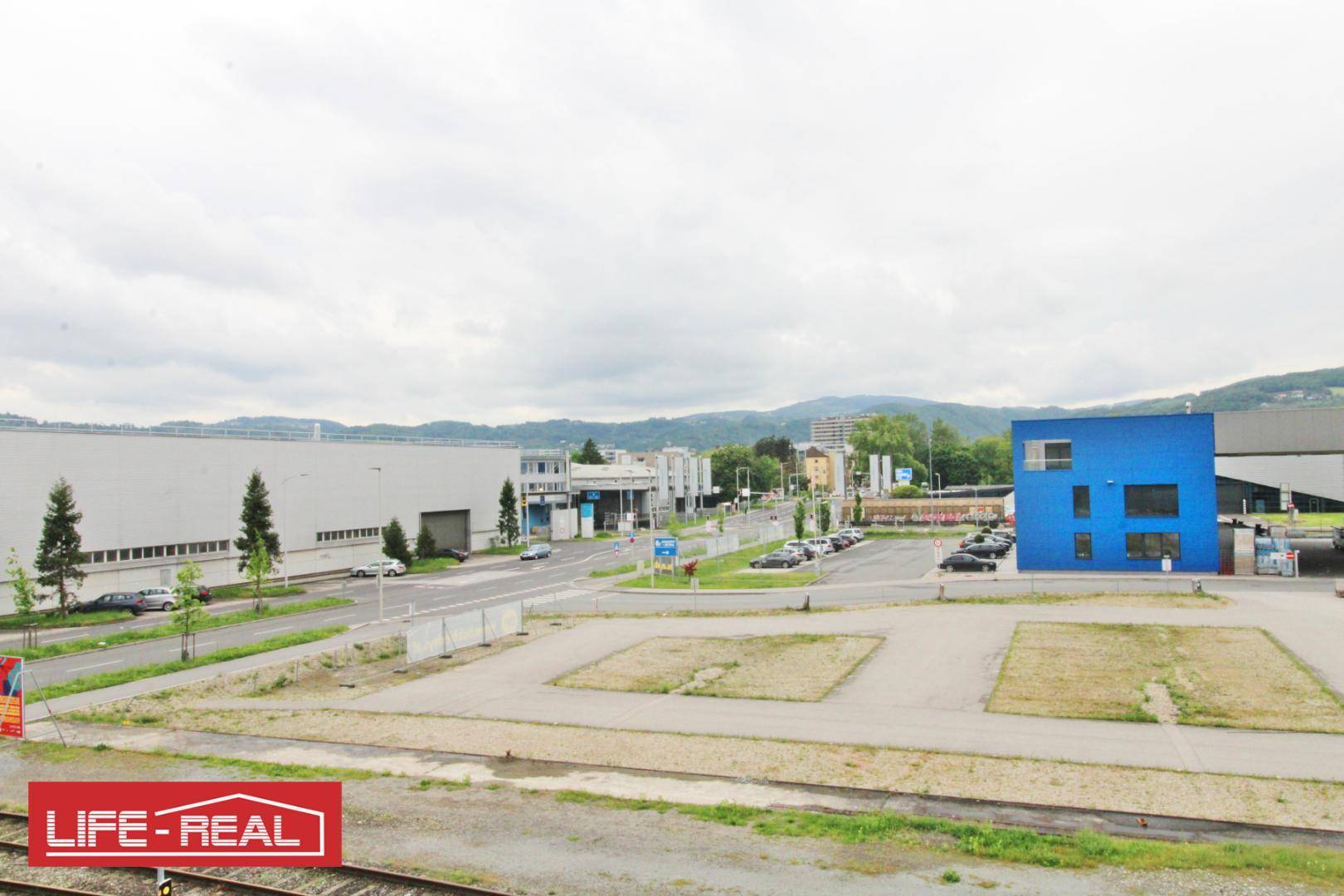 Industrielle Umgebung mit verschiedenen Gebäuden, Parkplätzen und einer Straße, umgeben von grünen Hügeln.