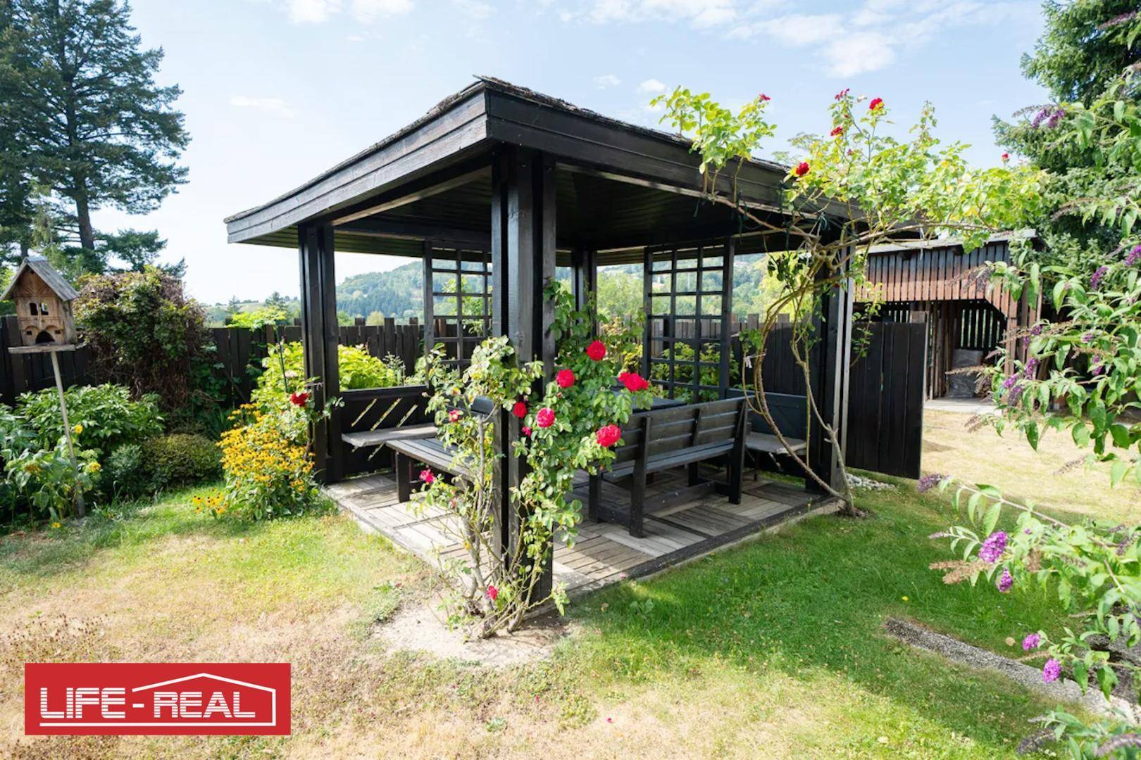 Gemütlicher Holzpavillon im Garten mit Sitzgelegenheiten, umrankt von blühenden Rosen.