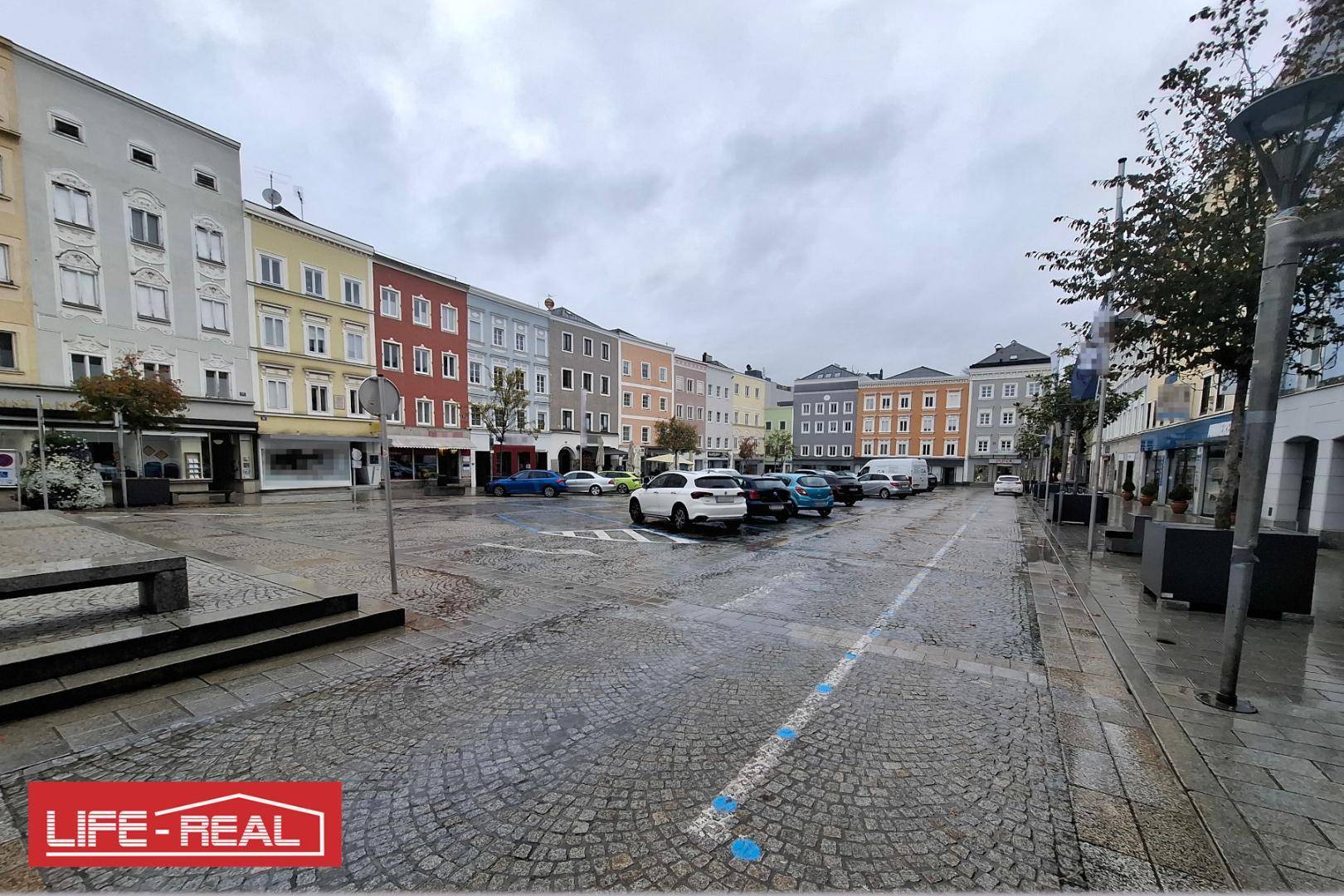 Blick über einen gepflasterten Stadtplatz mit parkenden Autos und farbenfrohen Altbauten.