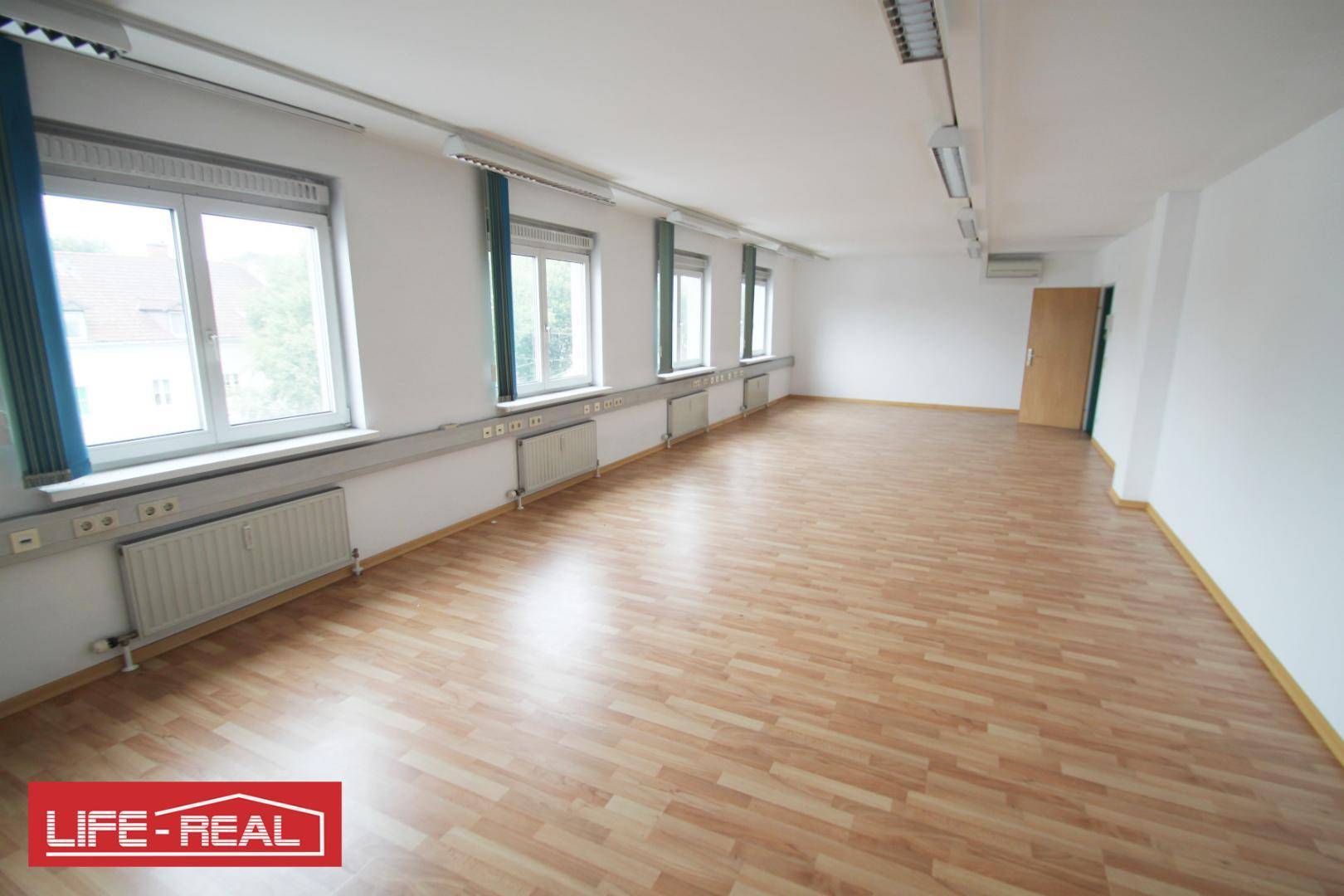 Heller Büroraum mit Laminatboden, weißen Wänden und einer Reihe von Fenstern, die den Raum belichten.