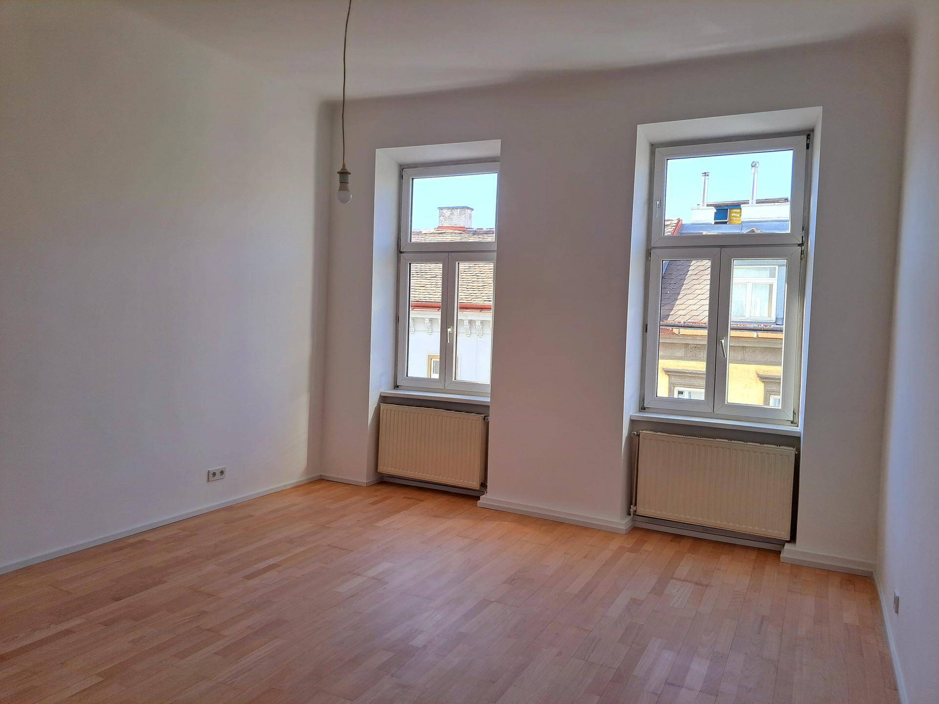 Leeres Zimmer mit Parkettboden und zwei Fenstern, die einen Blick auf die umliegenden Gebäude bieten.