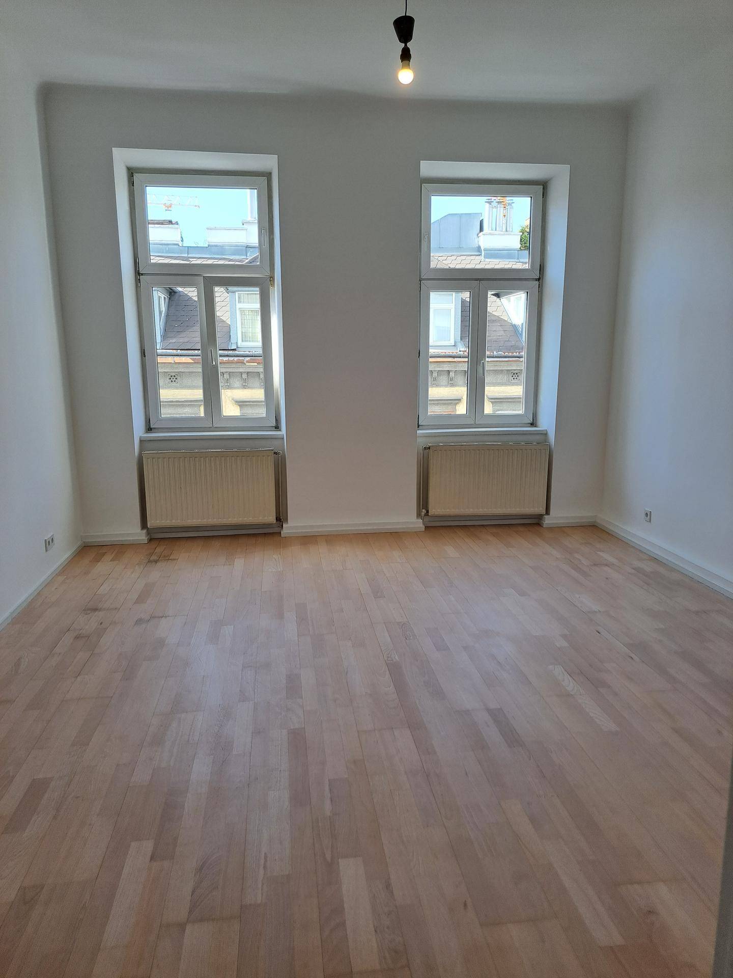 Geräumiges, unmöbliertes Zimmer mit hellem Parkettboden und zwei Fenstern, die viel Tageslicht hereinlassen.