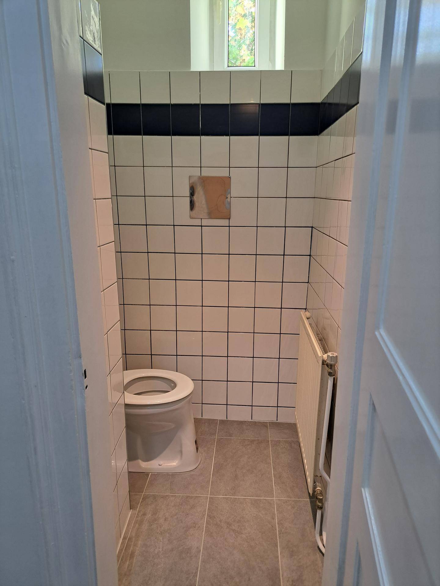 Separate Toilette mit weiß-grauen Fliesen und einem kleinen Fenster für Belüftung.