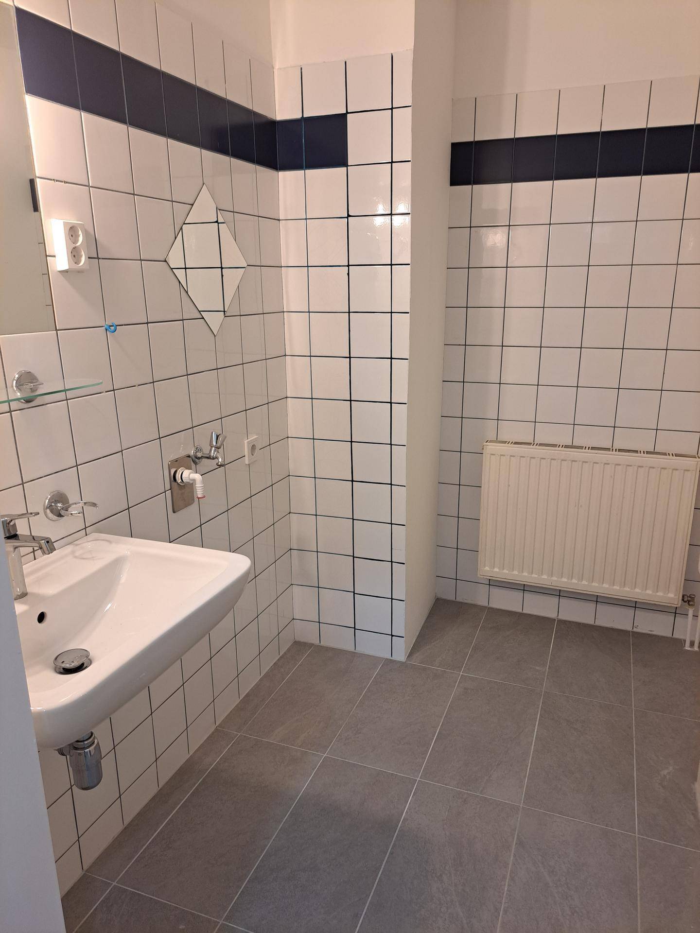 Badezimmer mit weiß-grauen Fliesen, einem Waschbecken und einem Spiegel, in gutem Zustand.