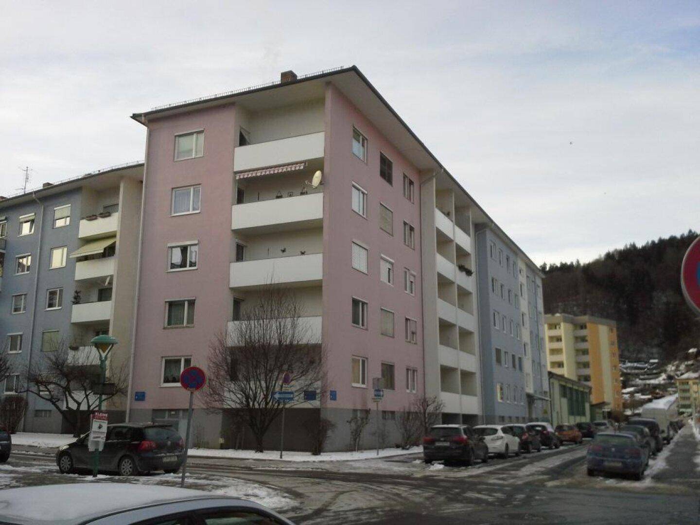 Mehrfamilienhaus mit rosa und blauer Fassade, Balkonen und umliegender Straße.