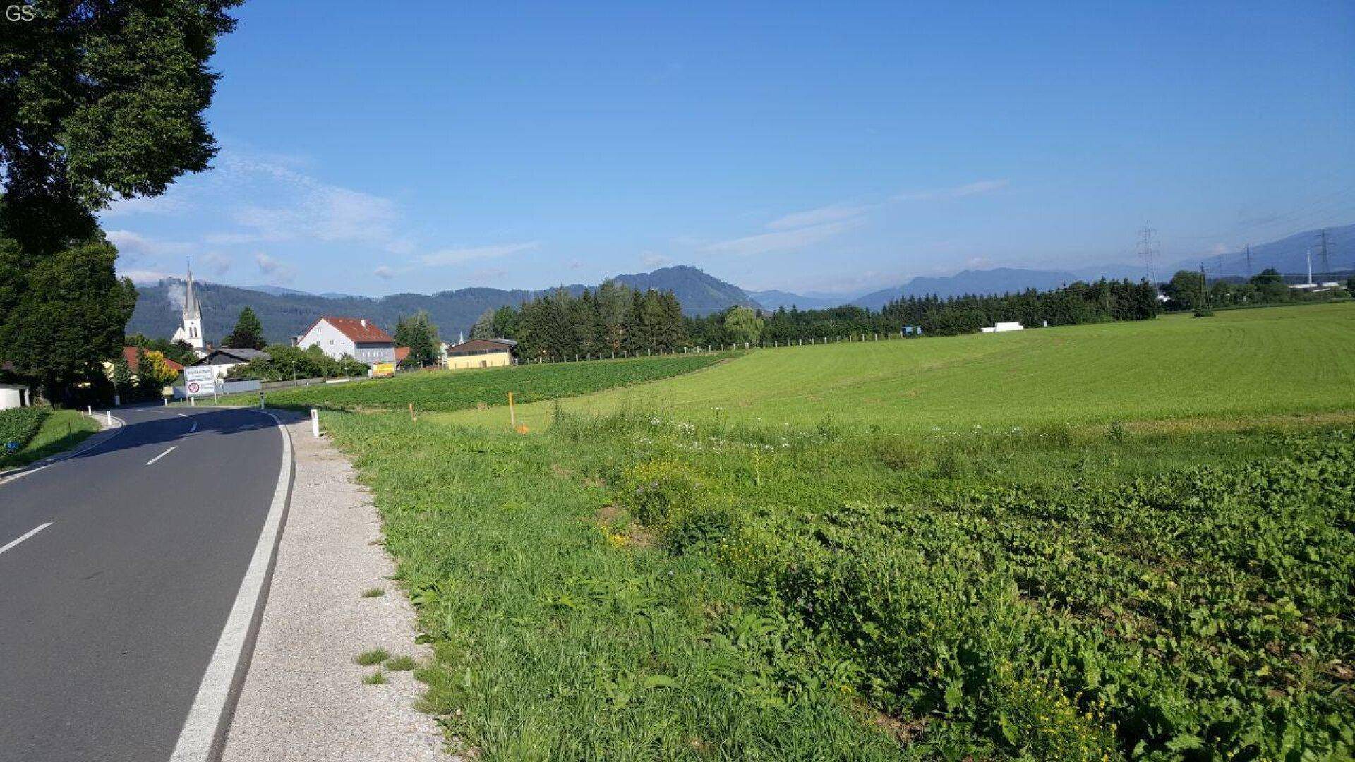Grüne Felder und eine Landstraße mit Häusern und Bergen in der Ferne unter klarem Himmel.