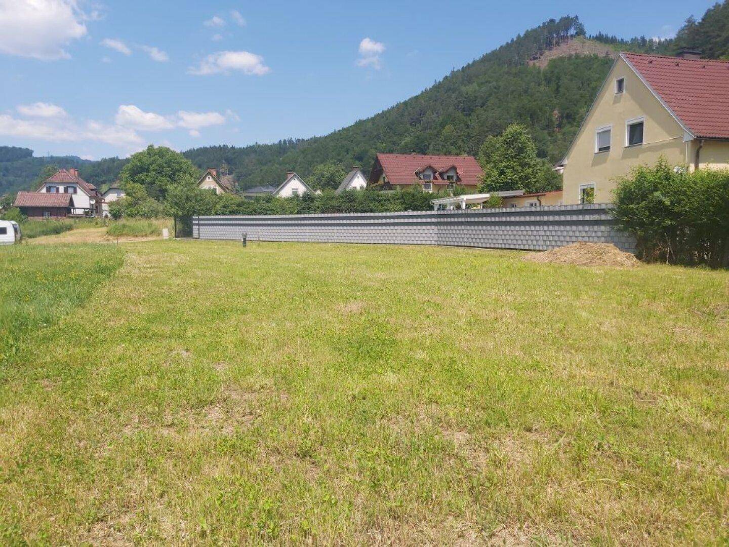 Sonniges Grundstück mit Rasenfläche, umgeben von Häusern und grünen Hügeln im Hintergrund.