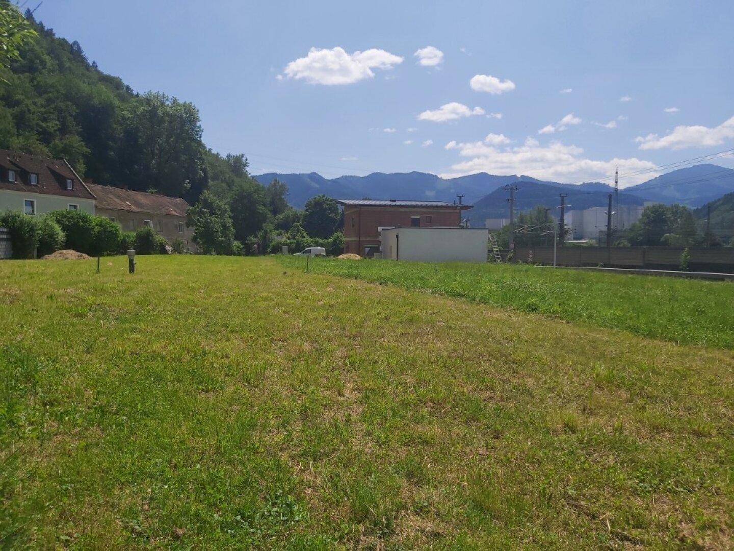 Weitläufiges grünes Grundstück mit Fernblick auf Berge und angrenzende Bebauung an einem sonnigen Tag.