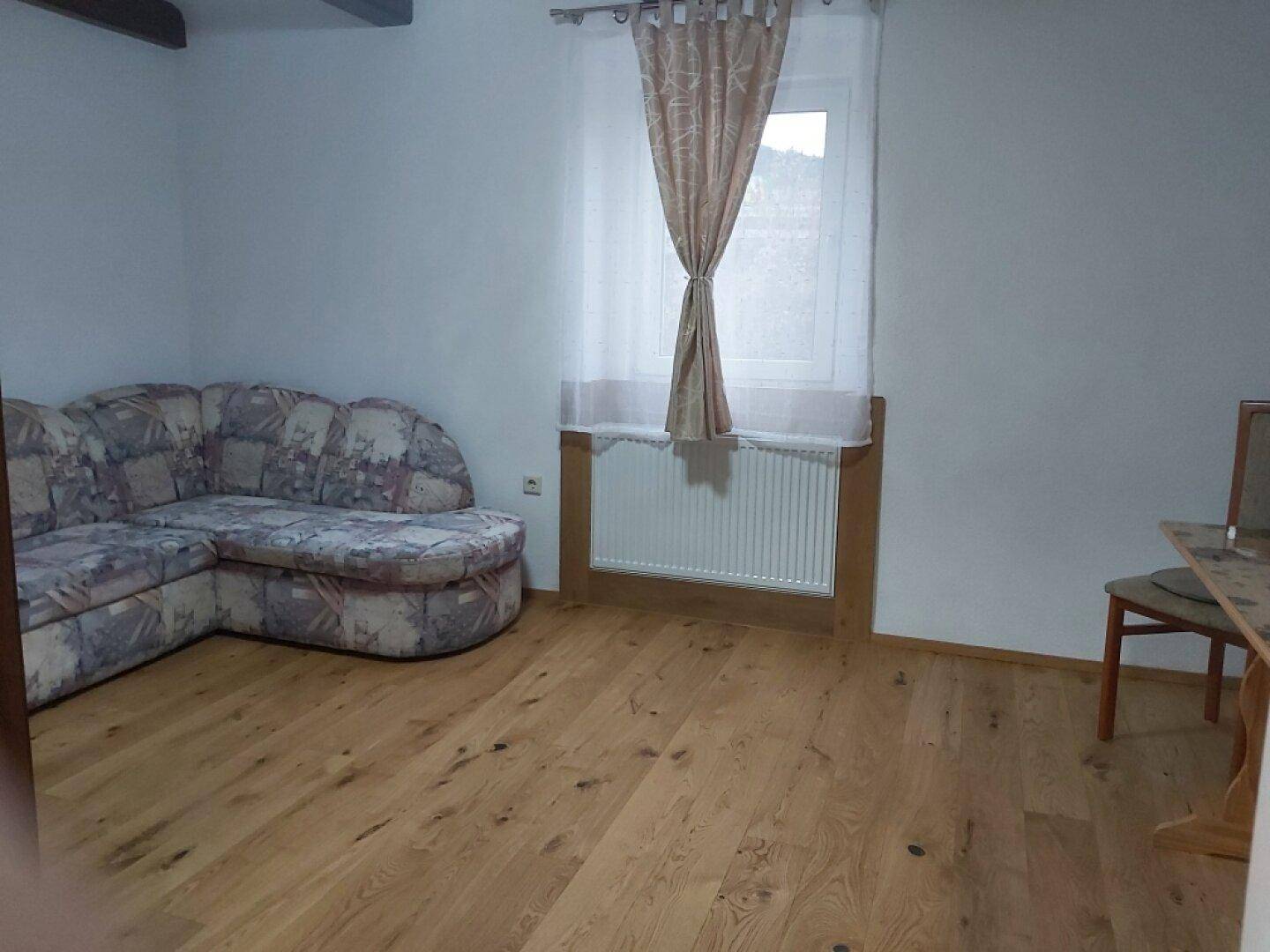 Wohnzimmer mit Ecksofa, Parkettboden und Fenster mit Vorhängen.