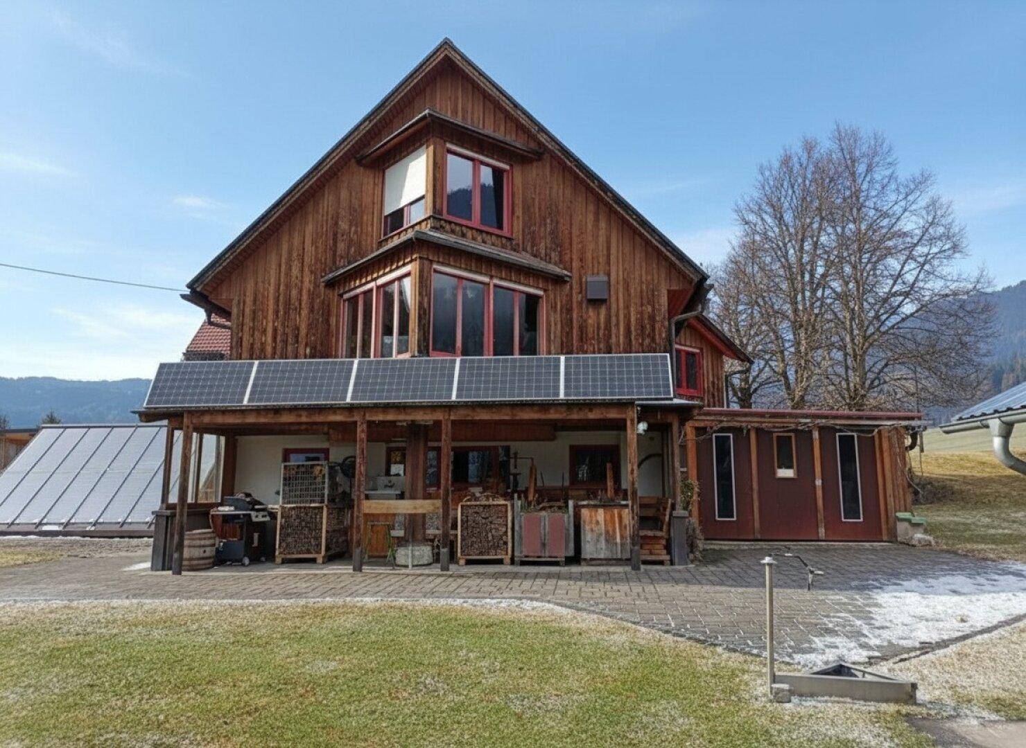 Rustikales Holzhaus mit Photovoltaikanlage auf dem Carport und Bergblick in ländlicher Umgebung.