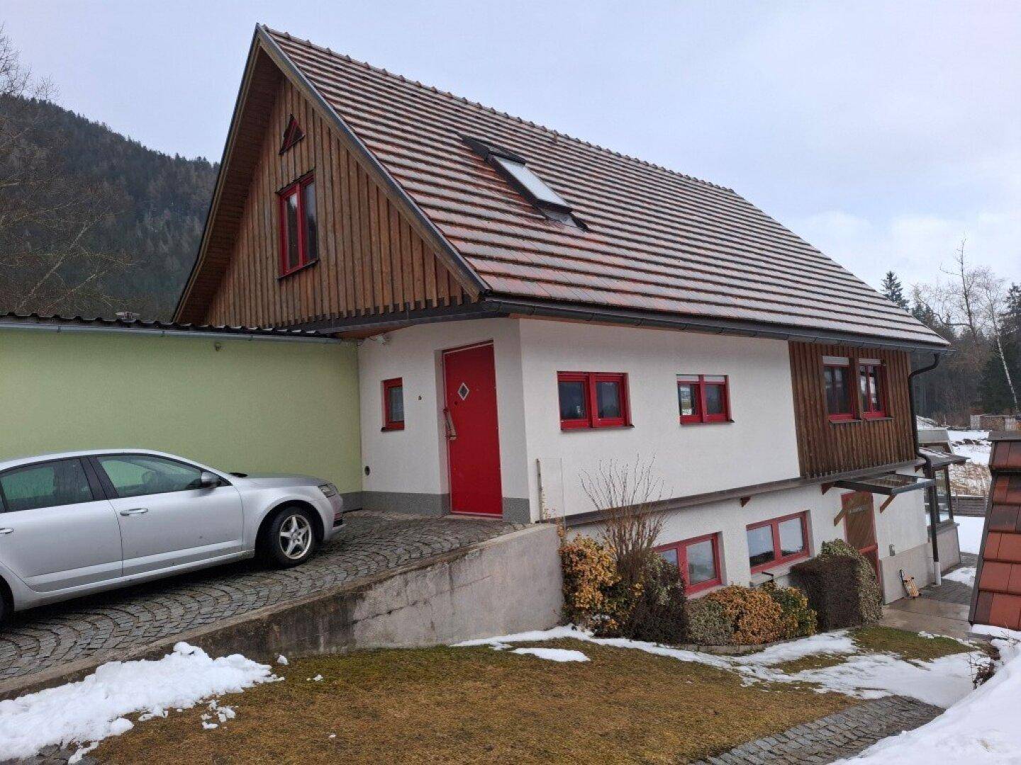 Seitenansicht des Hauses mit roter Eingangstür, Auffahrt und schneebedecktem Vorgarten.