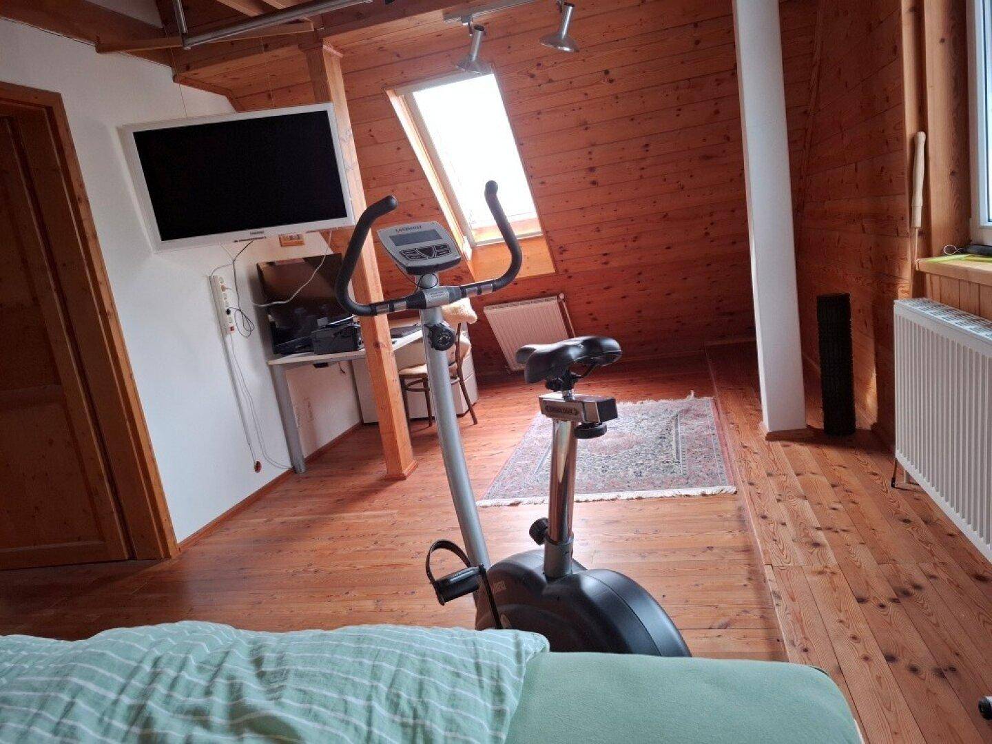 Dachgeschosszimmer mit Holzboden, Schrägdach und einem Heimtrainer für sportliche Aktivitäten.