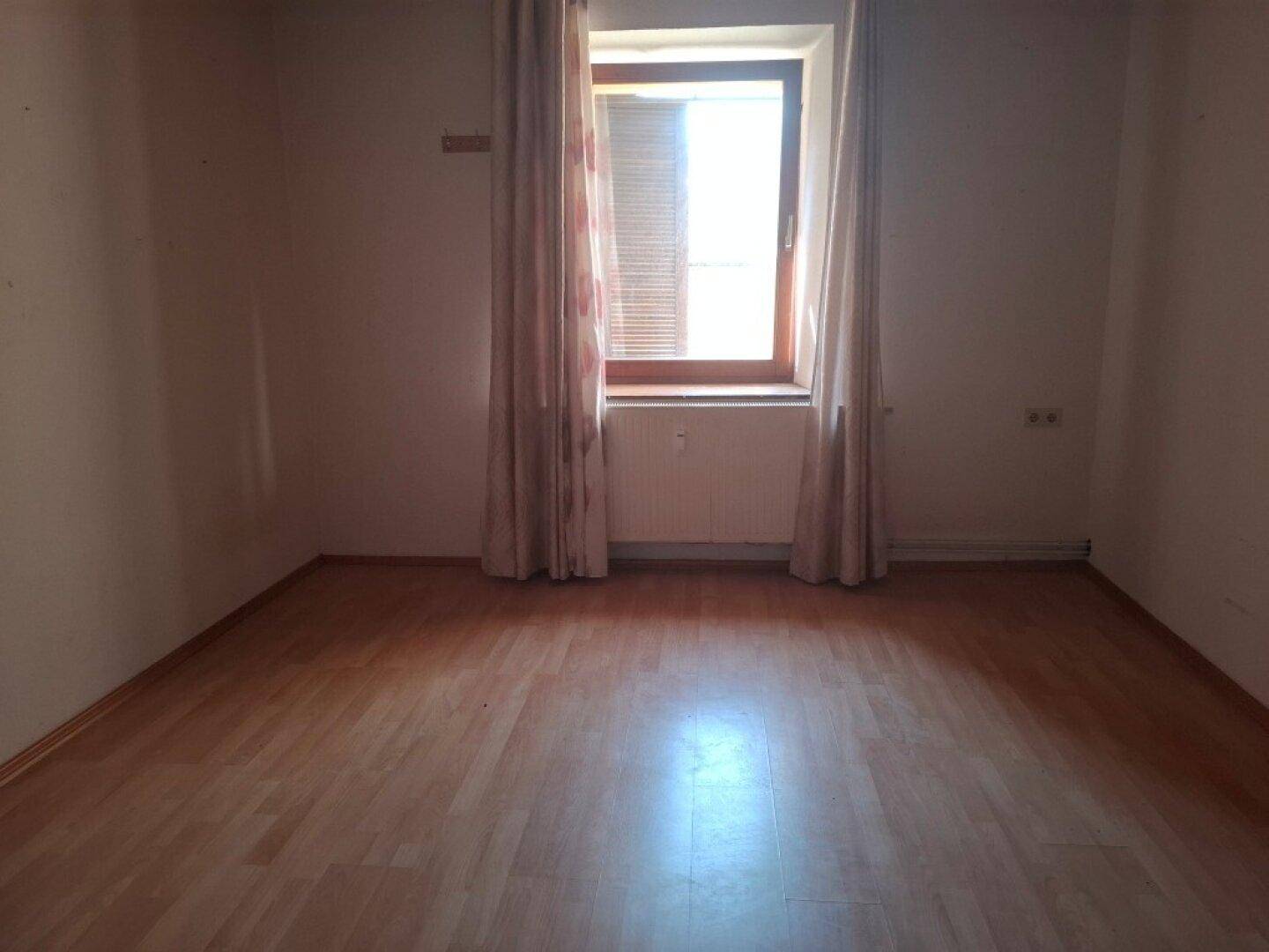 Leerer Raum mit Laminatboden, einem Fenster mit Jalousien und Vorhängen.