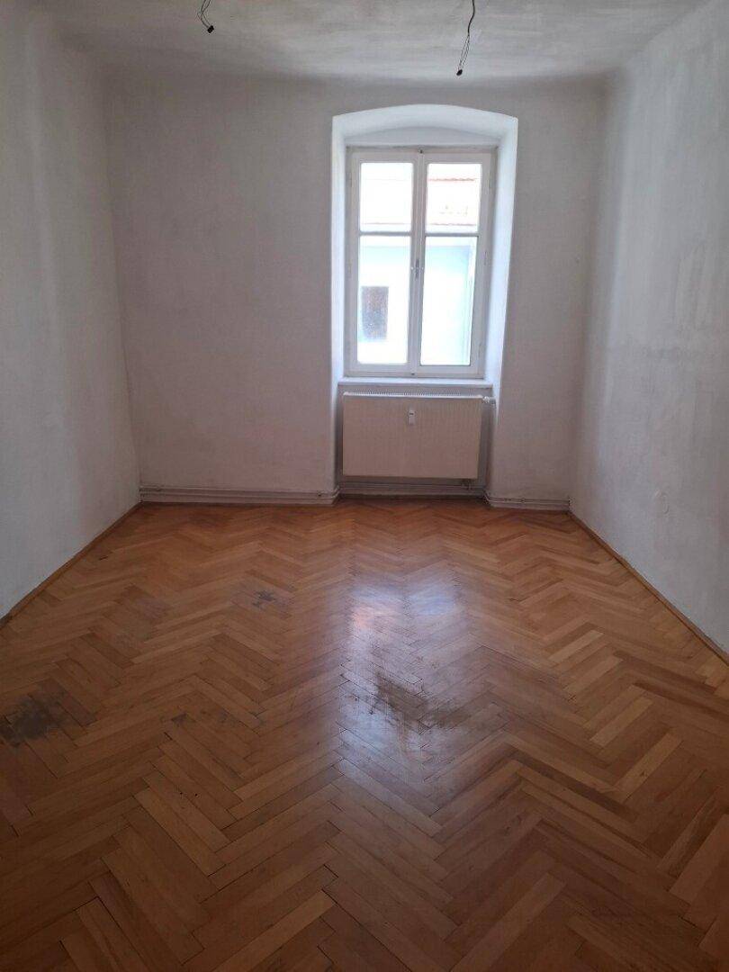 Leerer Raum mit Fischgrätparkettboden und einem Fenster, das Tageslicht hereinlässt.