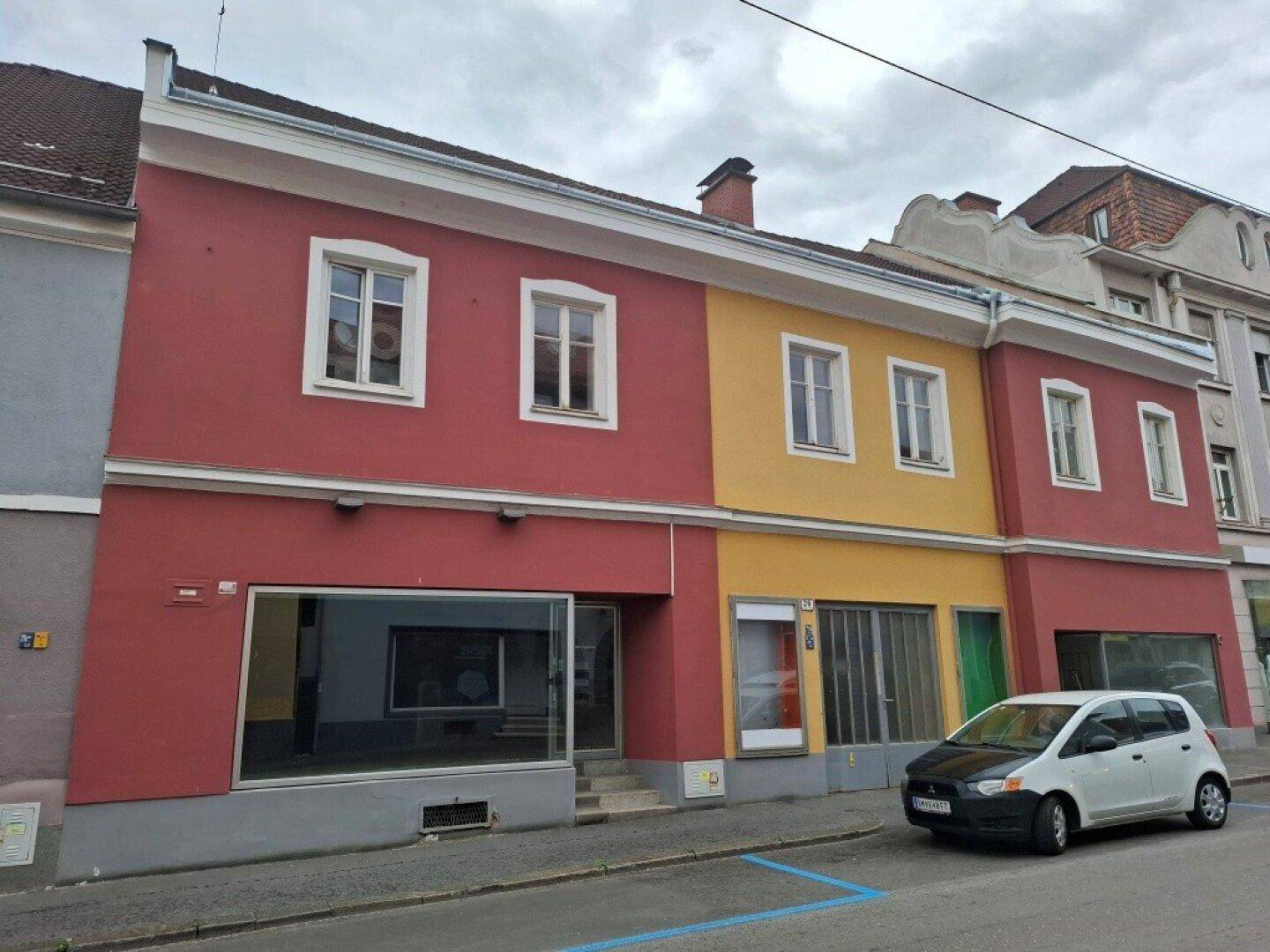 Fassade eines gemischt genutzten Gebäudes mit roten und gelben Abschnitten, großen Schaufenstern im Erdgeschoss und Wohnfenstern darüber.