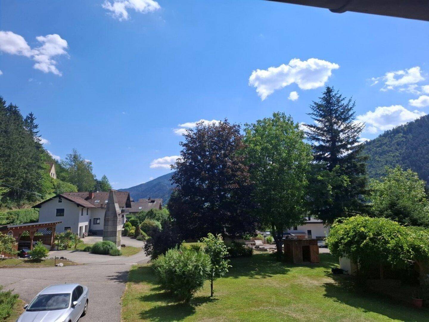 Weitläufiger Blick auf die umliegende Landschaft mit Häusern, Bäumen und Bergen unter blauem Himmel.