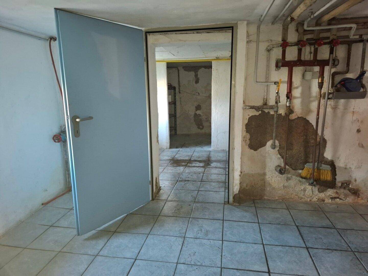 Kellerraum mit Fliesenboden, sichtbaren Rohren und Zugang zu einem weiteren Bereich.